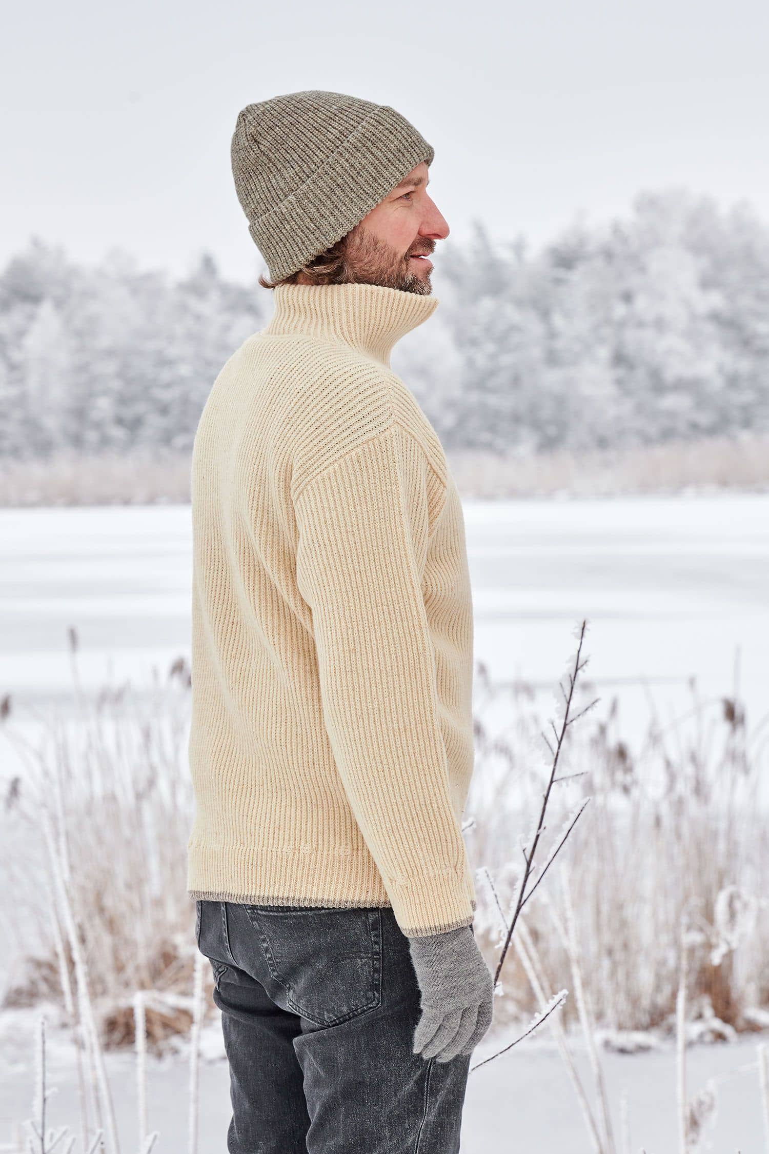 Mann in Schneelandschaft trögt einen naturweißen Finkhof Troyer aus bio Wolle (kbT), graue Handschuhe und Mütze
