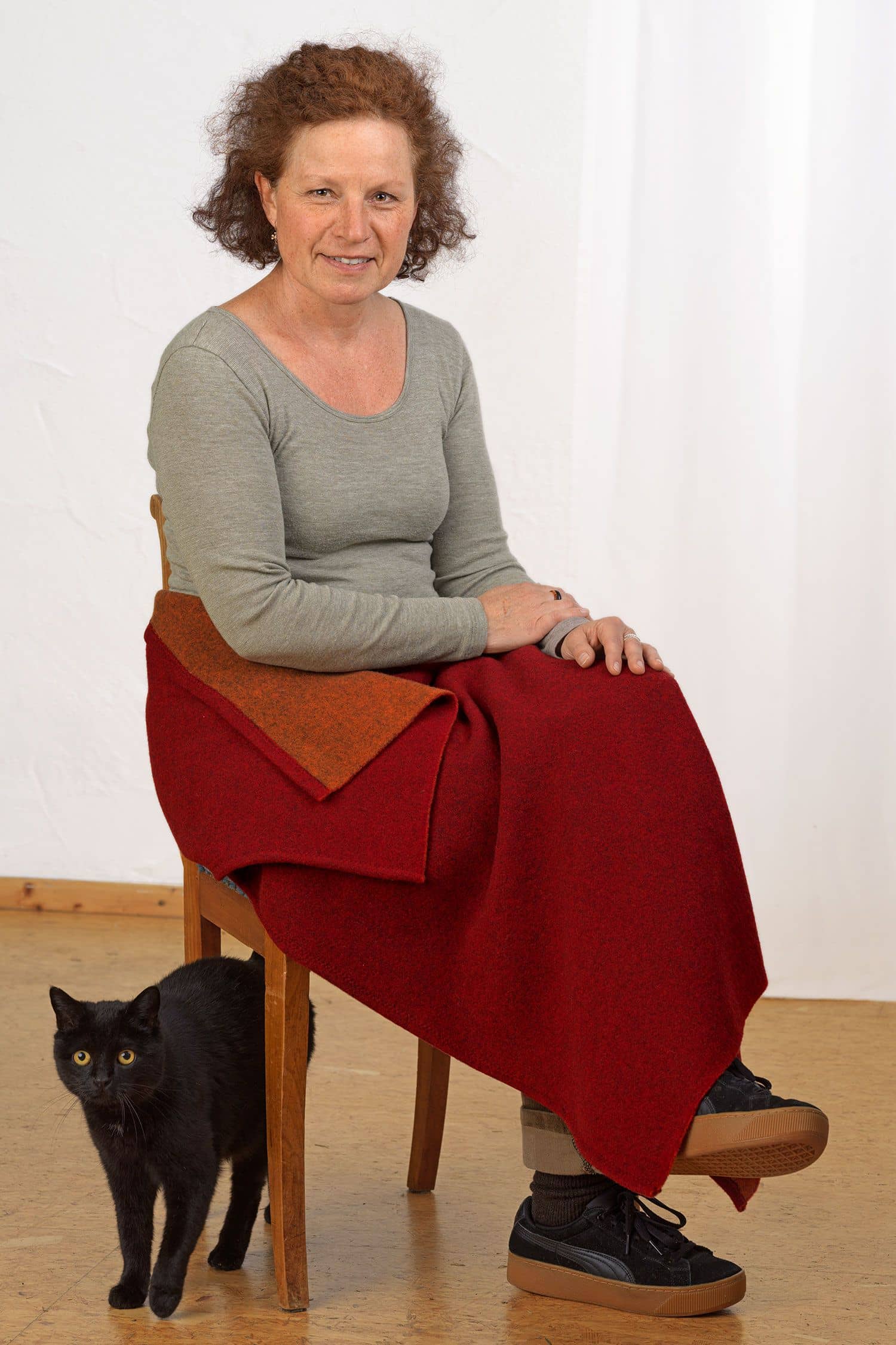 Frau sitzt mit roter Decke über den Beinen und einem unifarbigen Shirt auf einem Hocker, Katze steht daneben