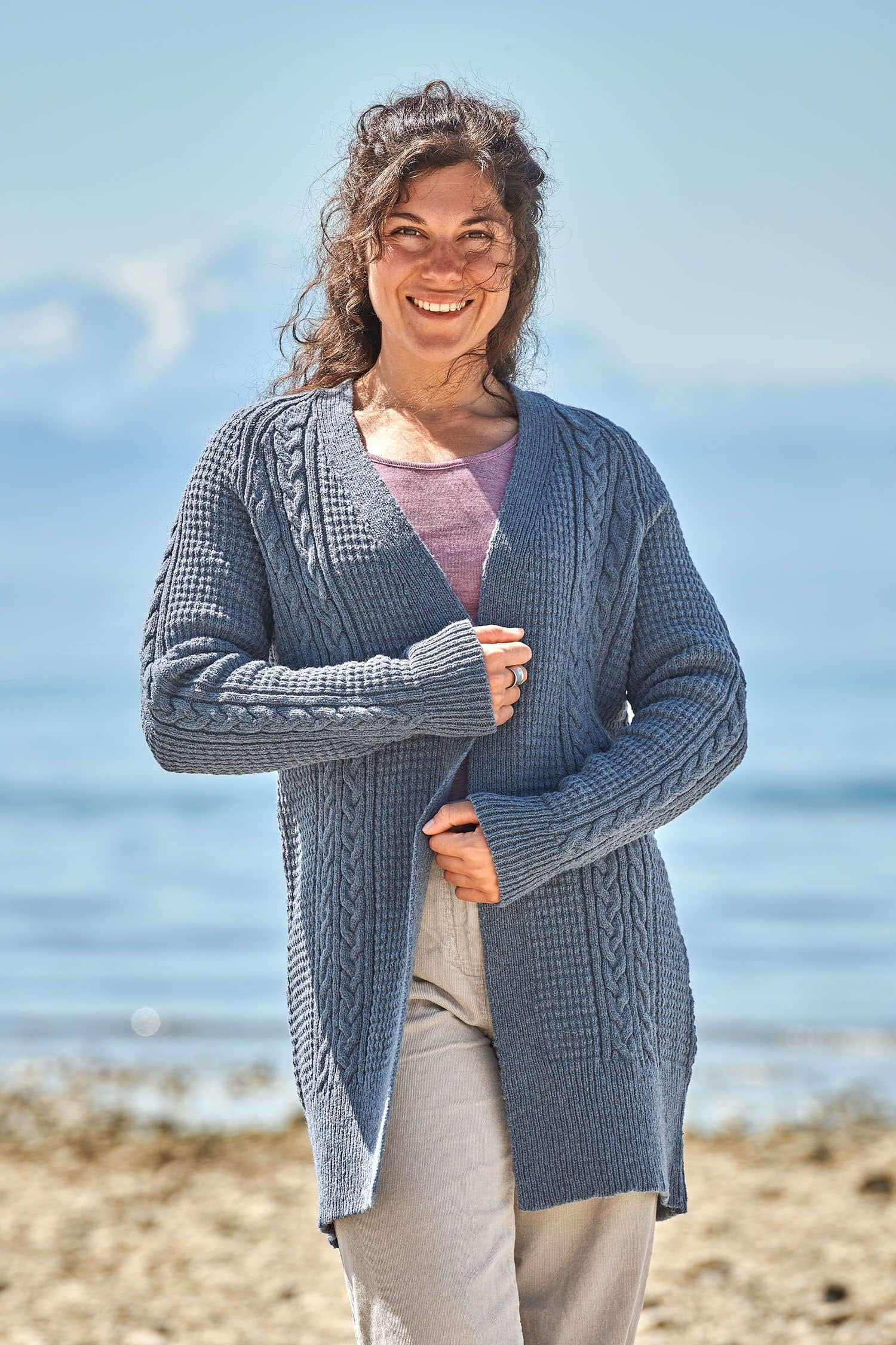 Frau trägt langen rauchblauen Cardigan mit Zopfmuster aus bio Wolle (kbT) vom Finkhof, eine beige Jeans und ein lila Shirt