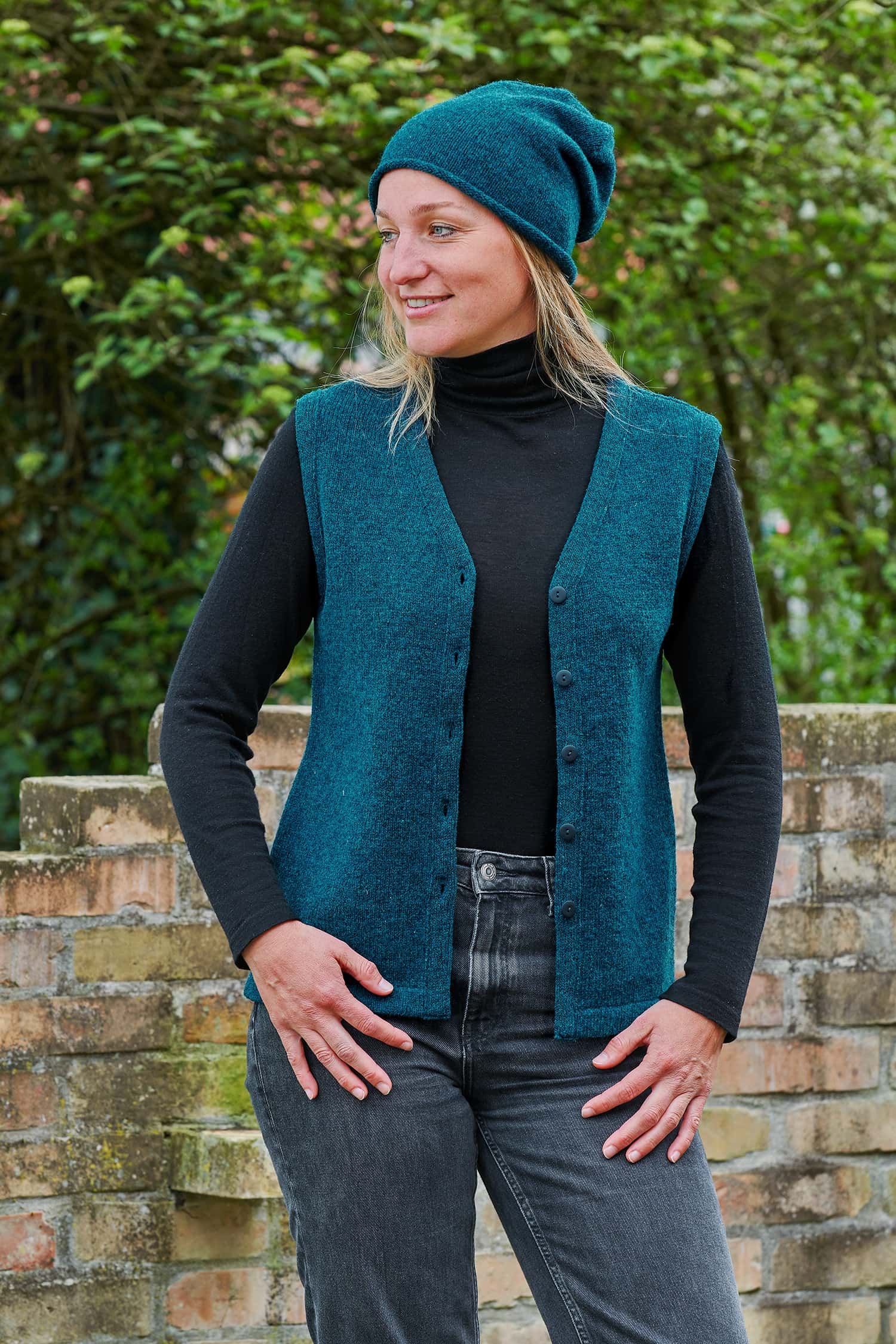Frau mit Jeans und schwarzem Shirt trägt eine türkise Mütze und eine feine türkise Strickweste vom Finkhof aus Bio-Wolle