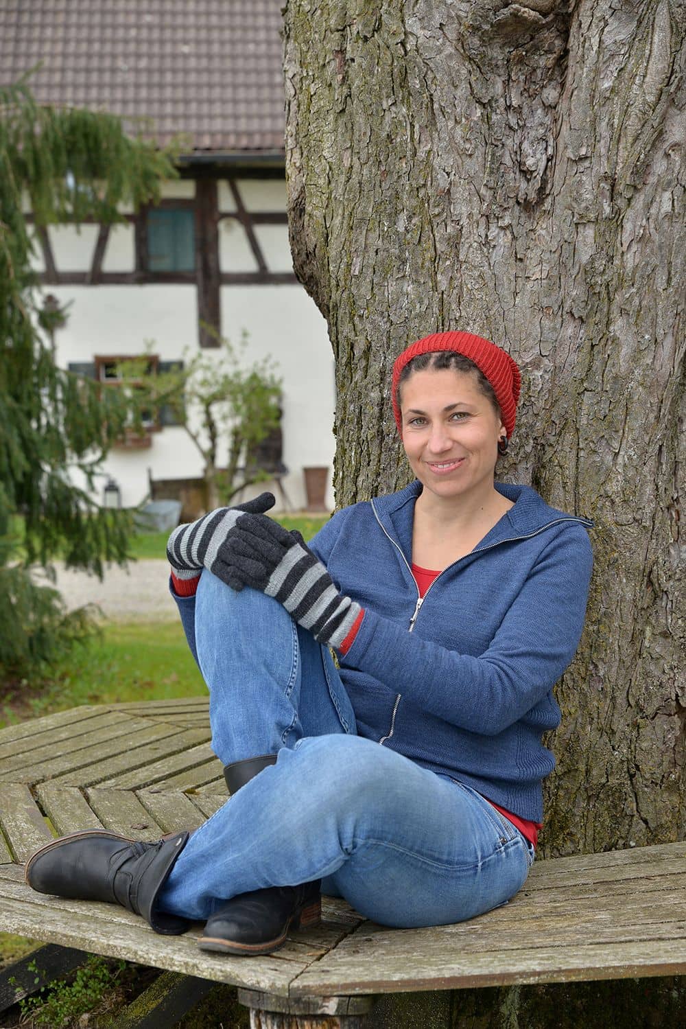 Frau auf Bank trägt eine Weste, Jeans, rote Mütze und geringelte Handschuhe in anthrazit/rot aus bio Finkhof Wolle (kbT)