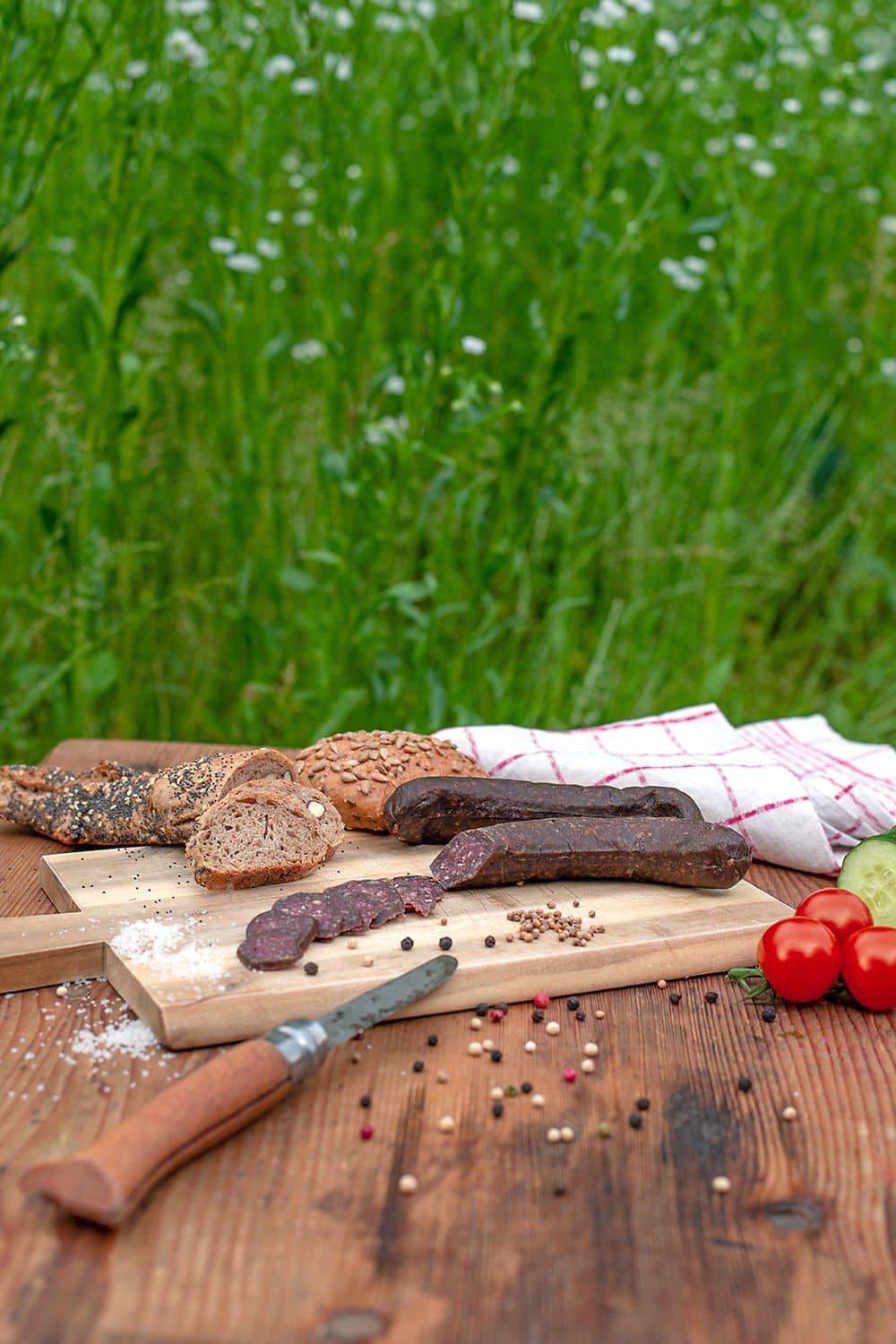 Bio Schafsalami vom Finkhof aufgeschnitten auf einem Brettchen mit Messer, Brot, Gemüse und Geschirrtuch daneben