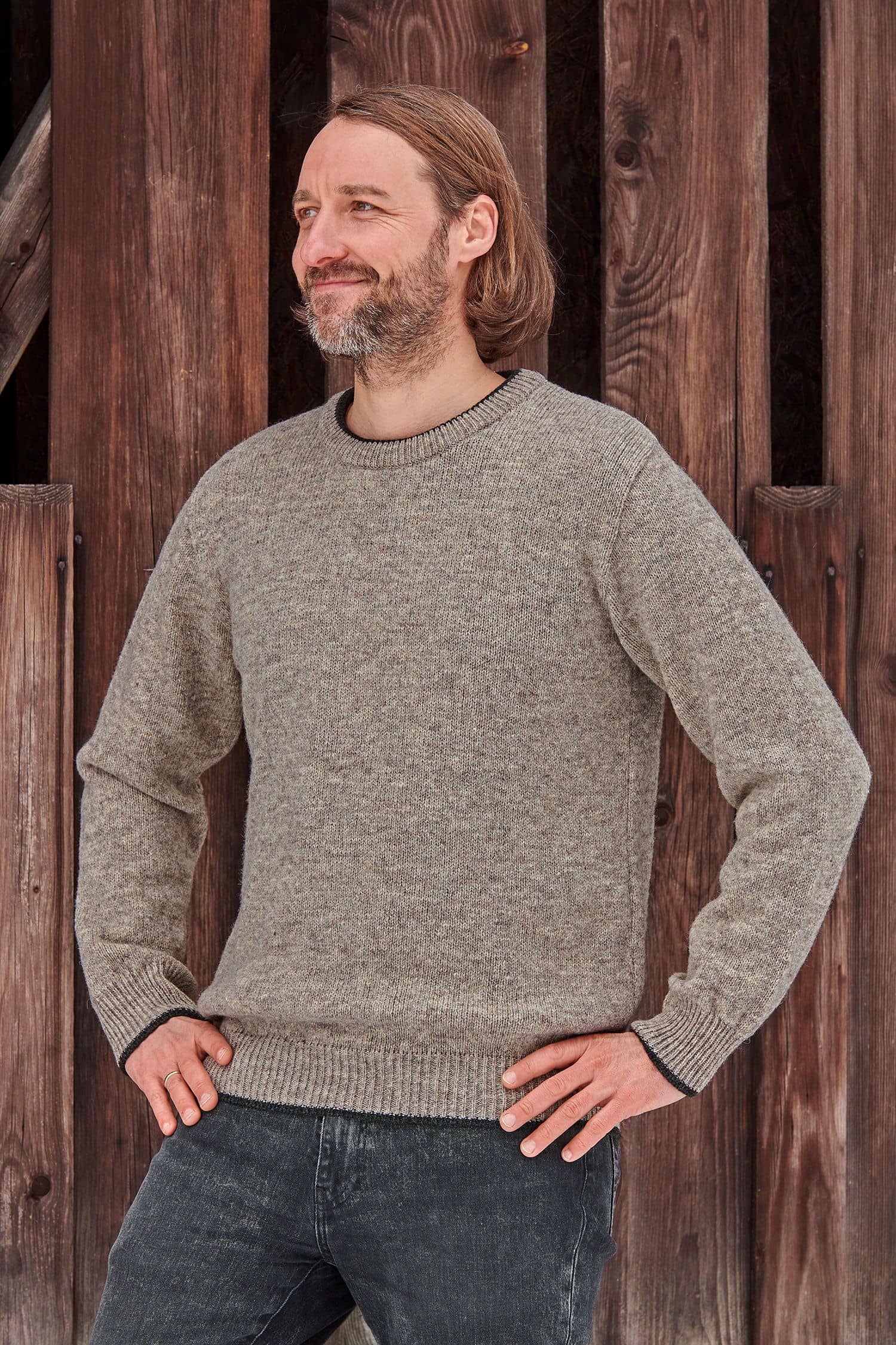 Mann steht mit grauem Finkhof Pulli aus bio Wolle (kbT) vor einer Holzfassade