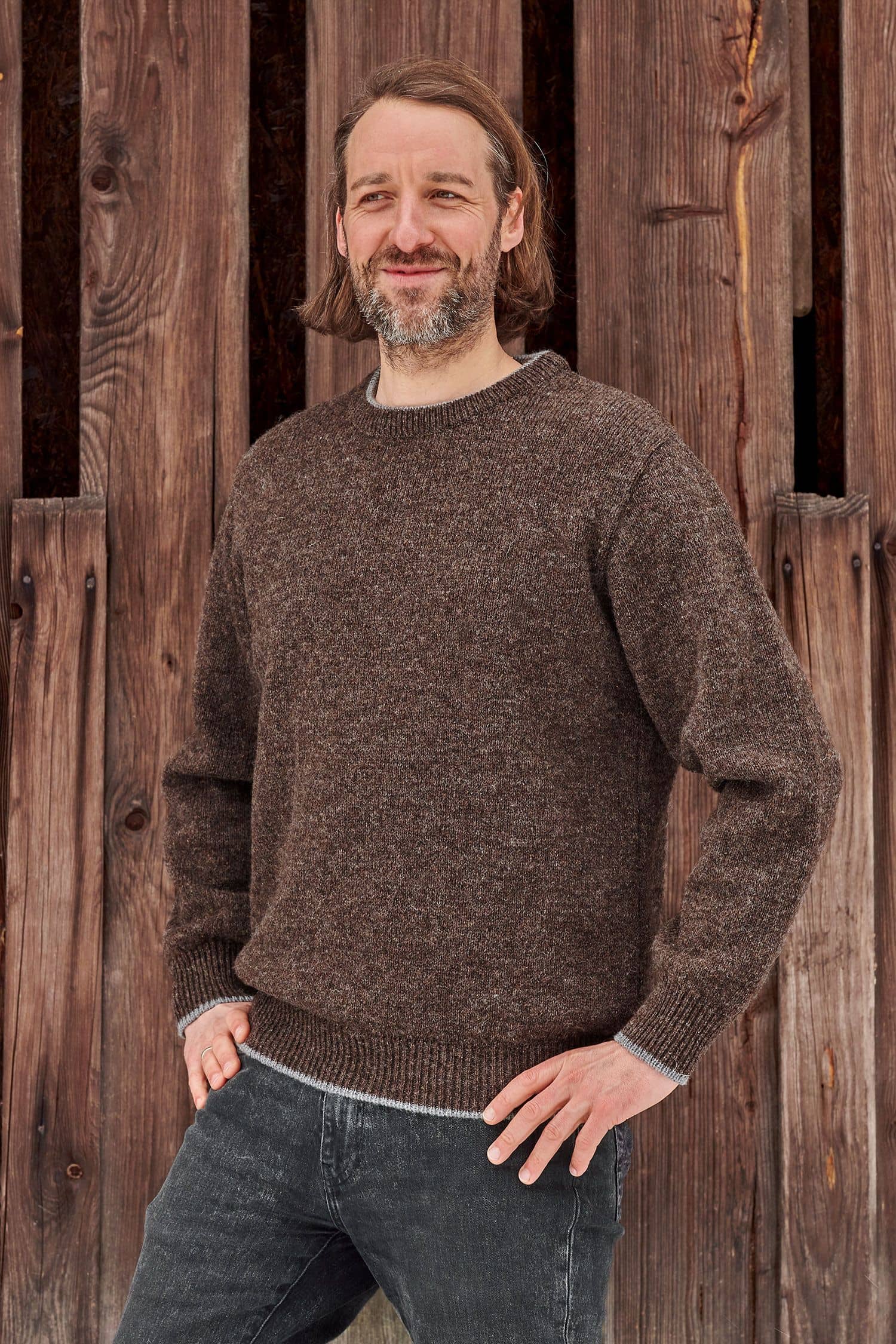 Mann steht mit braunem Finkhof Pulli aus bio Wolle (kbT) vor einer Holzfassade