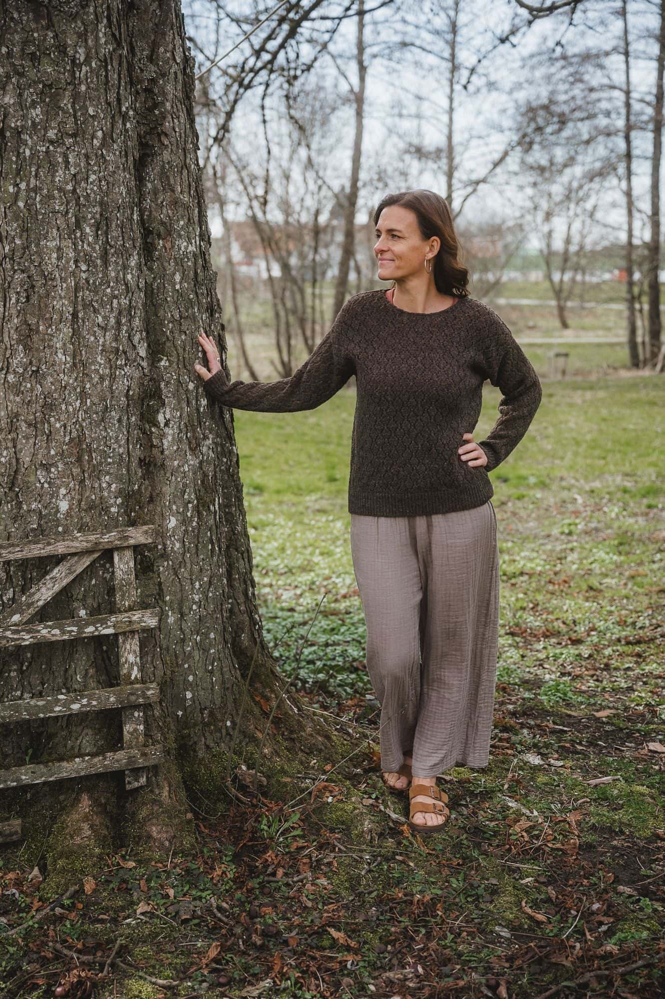 Finkhof-Ajour-Damen-Strickpullover ungefärbt braun Gr. S Frau steht neben Baum und trägt eine weite Hose mit braunem Strickpullover mit Ajourmuster aus Bio-Wolle vom Finkhof