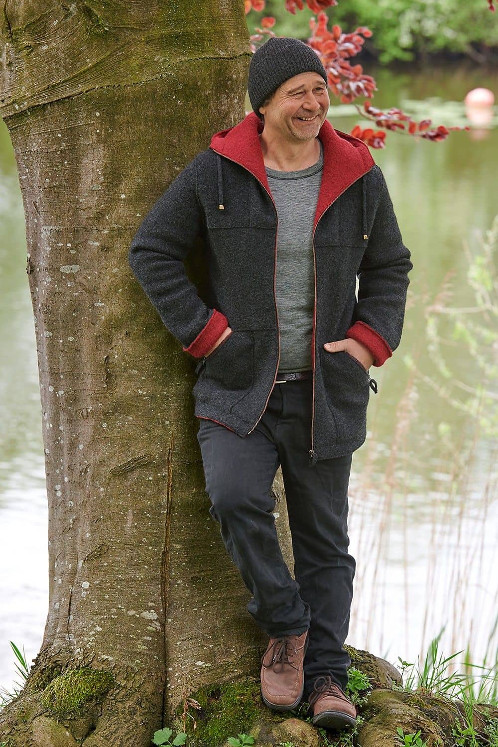 Mann lehnt an Baum und trägt Mütze, graues Shirt und dunkelgraue Jacke aus bio Wolle (kbT) vom Finkhof mit Kapuze
