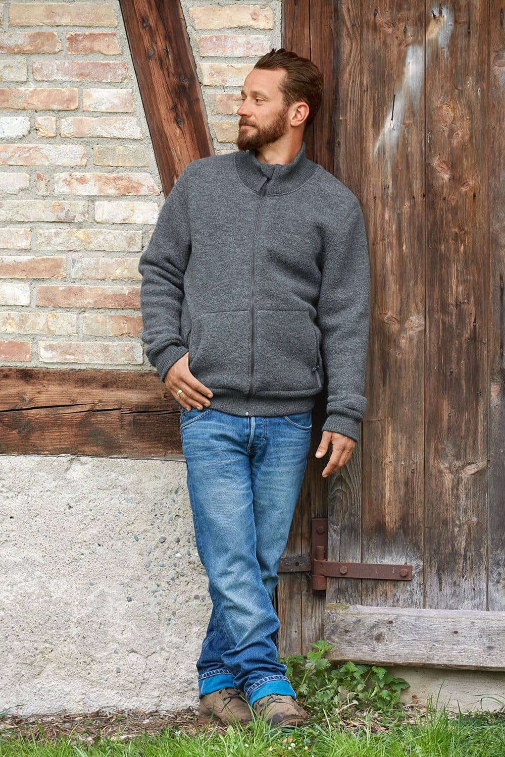 Mann vor Holztor trägt graue Finkhof Jacke Toni aus bio Wolle (kbT) mit Jeans und braunen Stiefeln