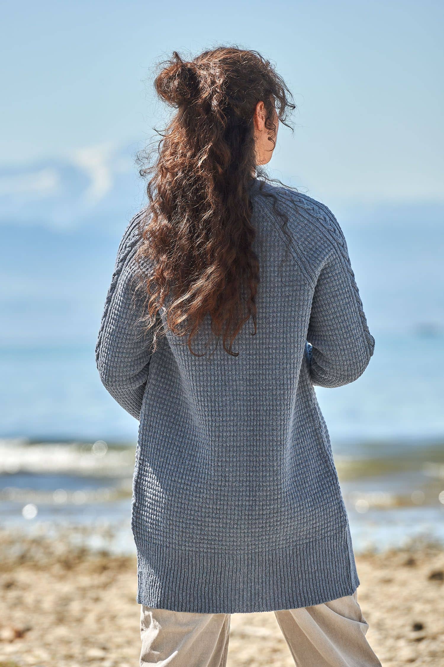 Rückansicht von Frau mit rauchblauem Cardigan mit Zopfmuster aus bio Wolle (kbT) vom Finkhof und beiger Jeans