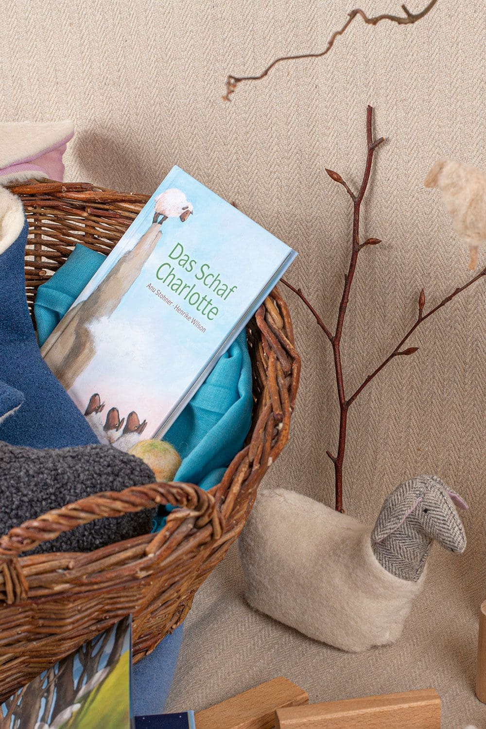 Buch ,,Das Schaf Charlotte" liegt in Korb mit Stoffen, Stoffschaf steht daneben und Äste im Hintergrund