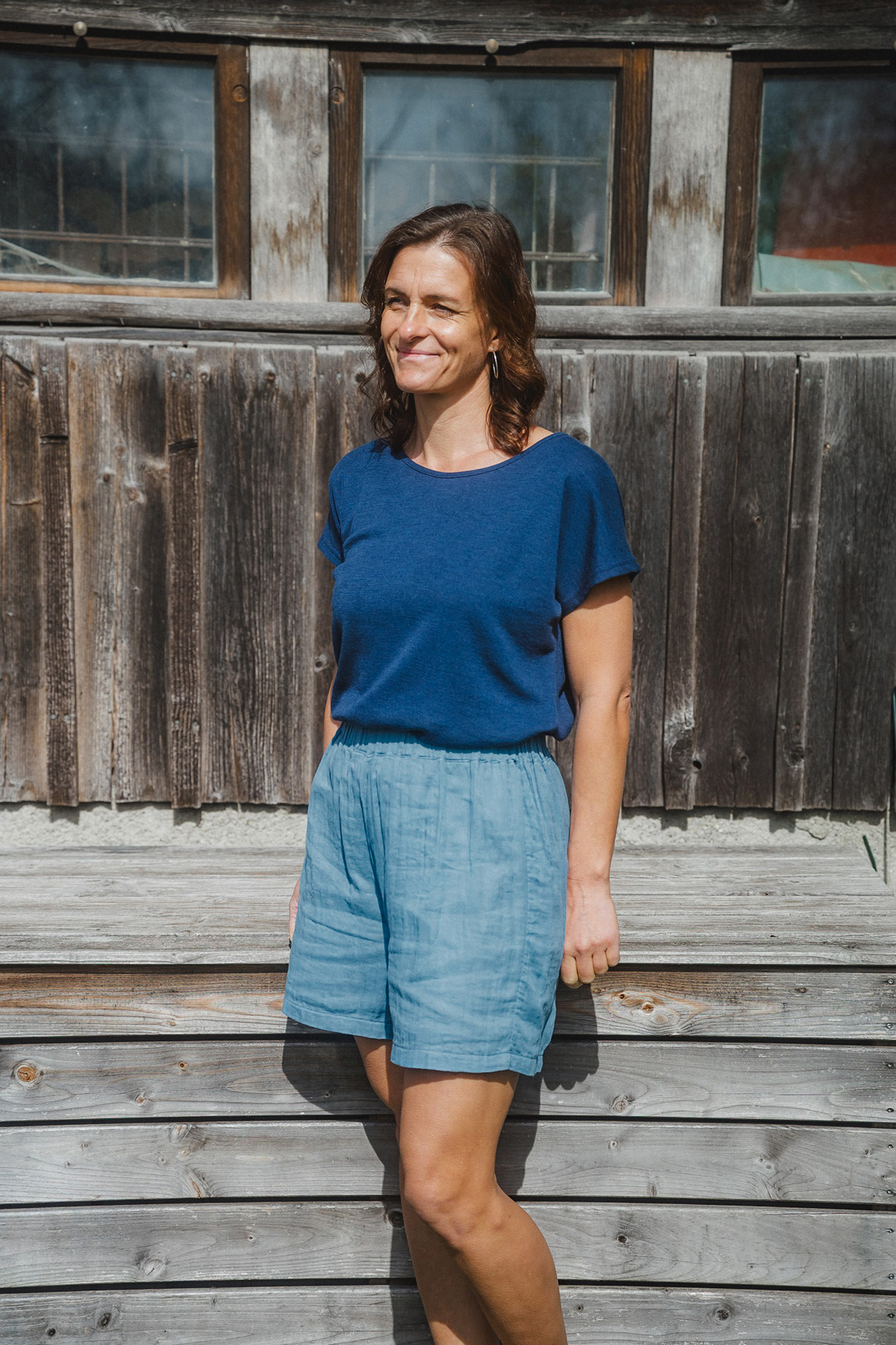 Damenshorts aus Bio-Baumwoll-Musselin Frau steht vor Holzwand und trägt ein blaues Shirt und eine blaue Shorts aus Bio-Baumwoll-Musselin (kbA) vom Finkhof
