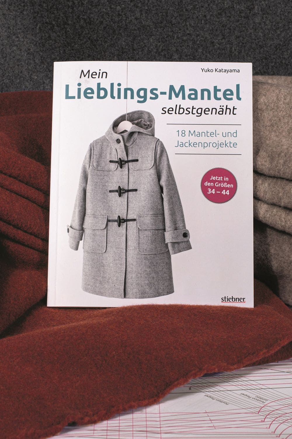 Mein Lieblings-Mantel selbstgenäht Buch ,,Mein Lieblingsmantel selbstgenäht" auf rotem Stoff und Stoffen im Hintergrund