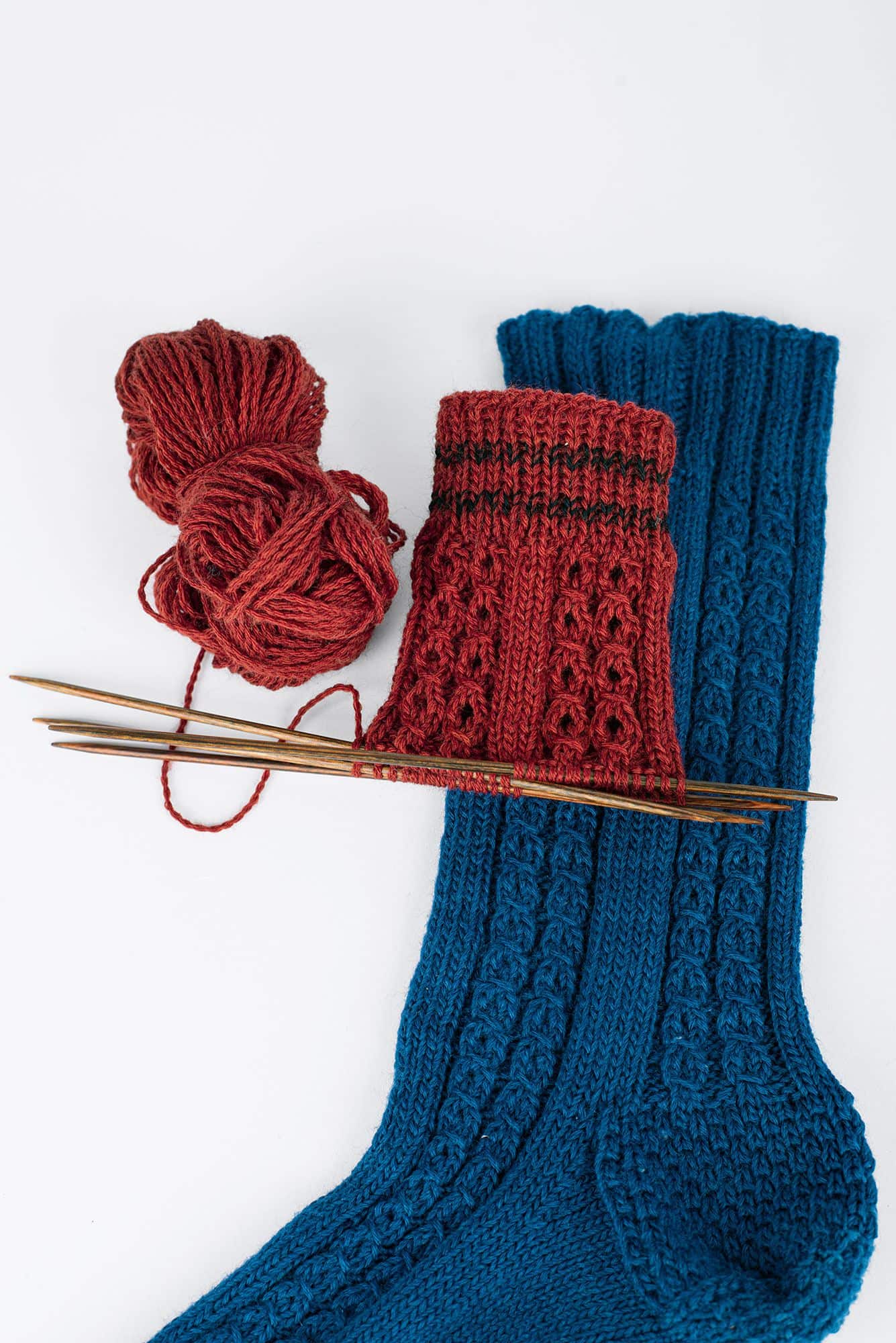 blaue Stricksocke und rote angefangene Stricksocke aus feiner Finkhof Sockenwolle auf weißem Untergrund