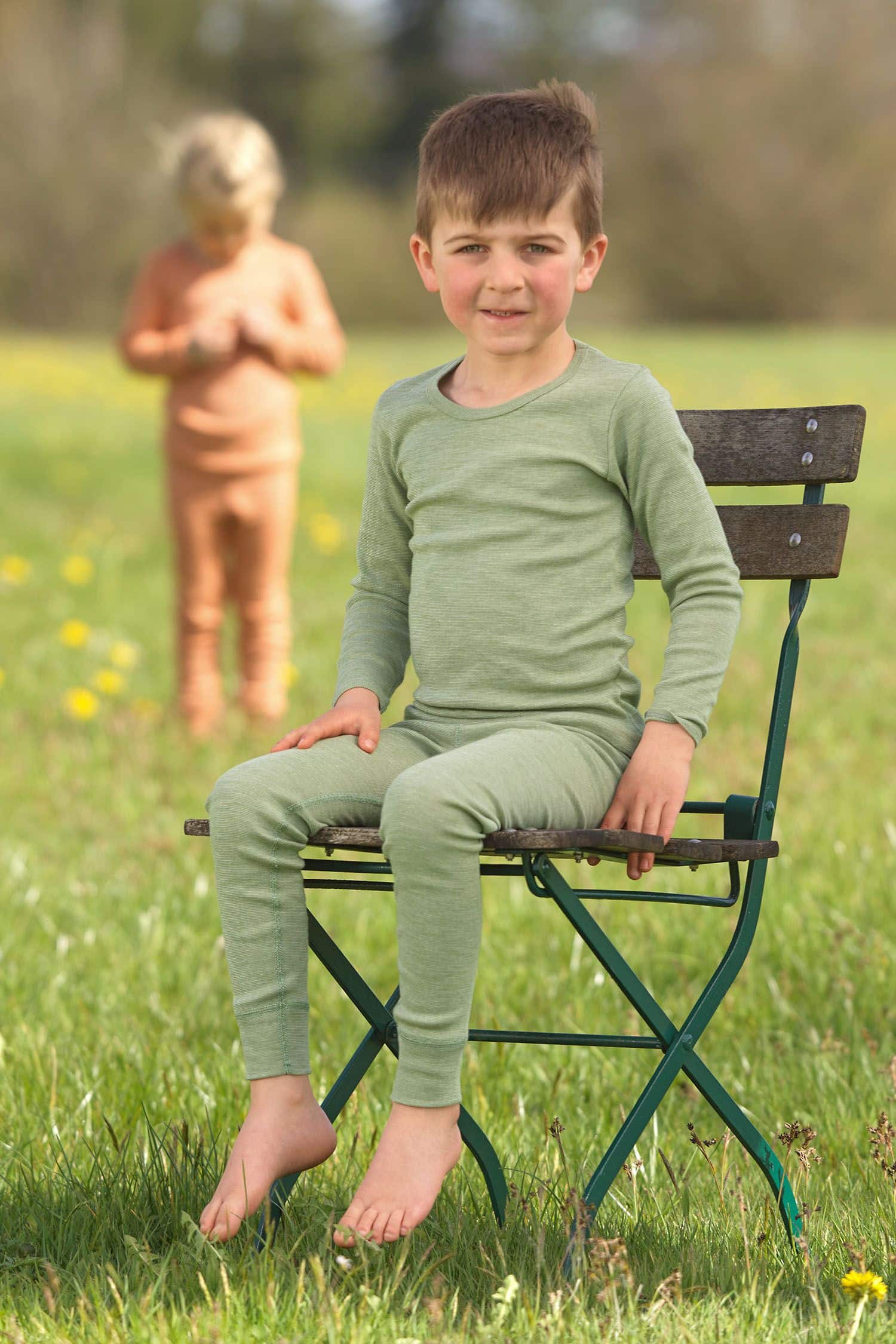 Kind sitzt auf Stuhl trägt ein grünes Langarmshirt aus bio Wolle (kbT), bio Baumwolle (kbA) und Seide mit langer Unterhose