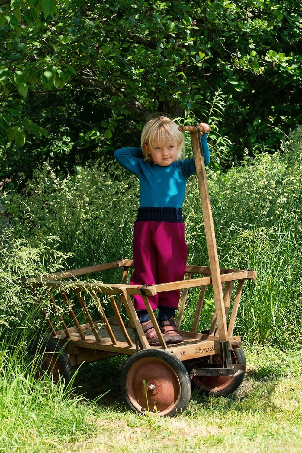 Kind steht in einem Leiterwagen und trägt ein blaues Shirt und eine pinke Walkhose aus bio Wolle (kbT) vom Finkhof