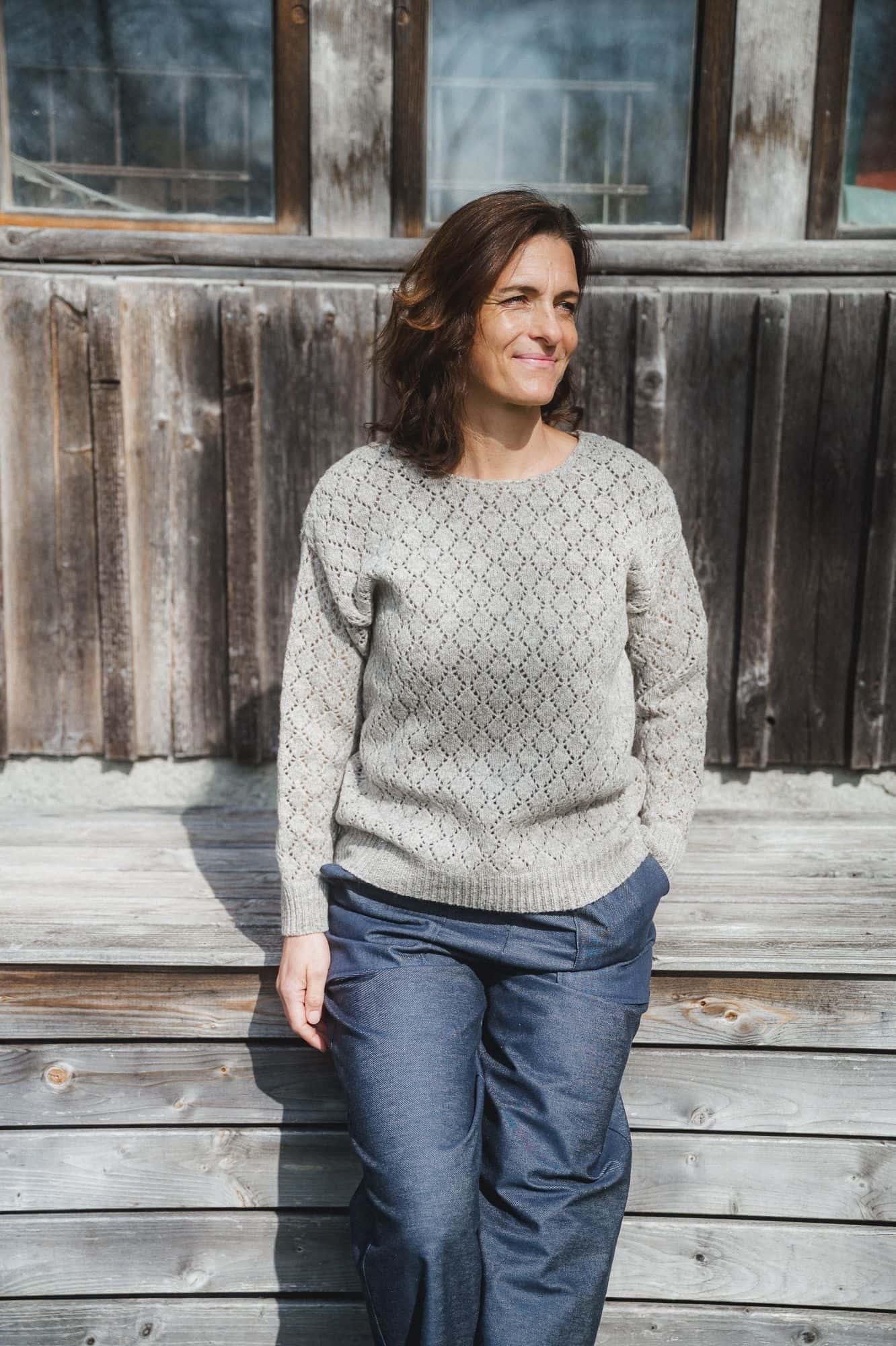 Finkhof-Ajour-Damen-Strickpullover ungefärbt grau Gr. S Frau steht vor Holzwand und trägt grauen Strickpullover mit Ajourmuster aus Bio-Wolle vom Finkhof