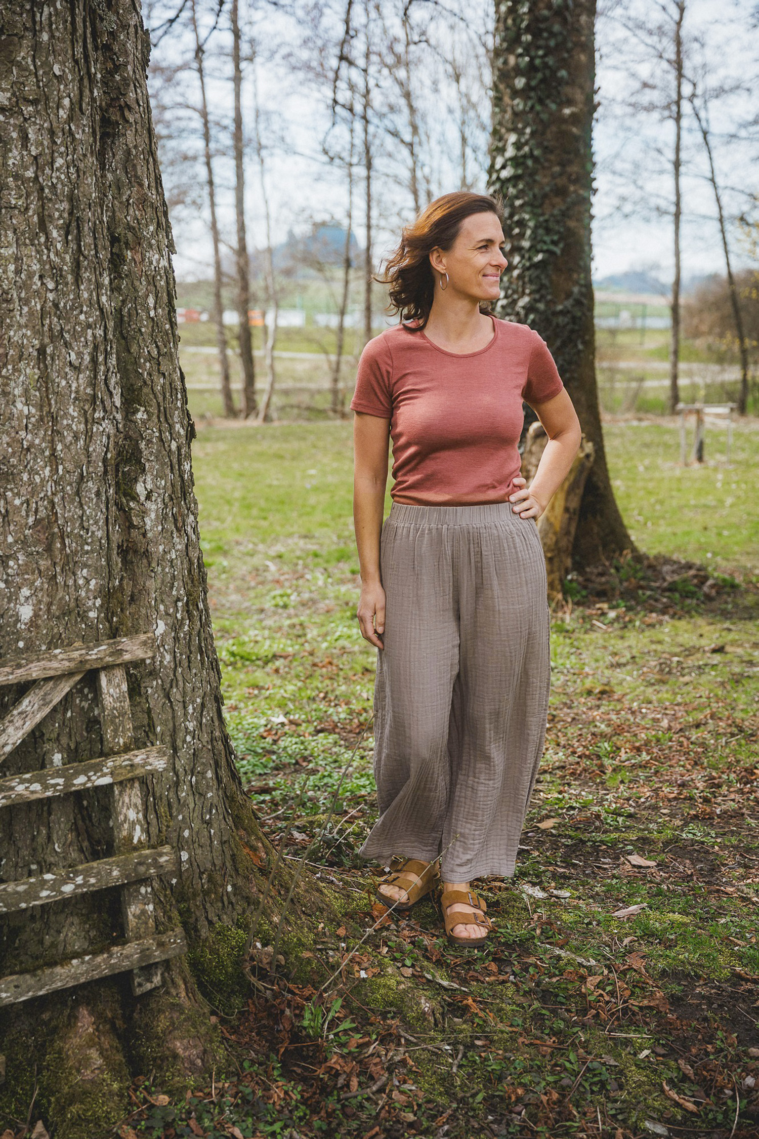 Damenhose aus Bio-Baumwoll-Musselin Frau steht neben Baum und trägt Shirt und braune weite Hose aus Bio-Baumwoll-Musselin vom Finkhof