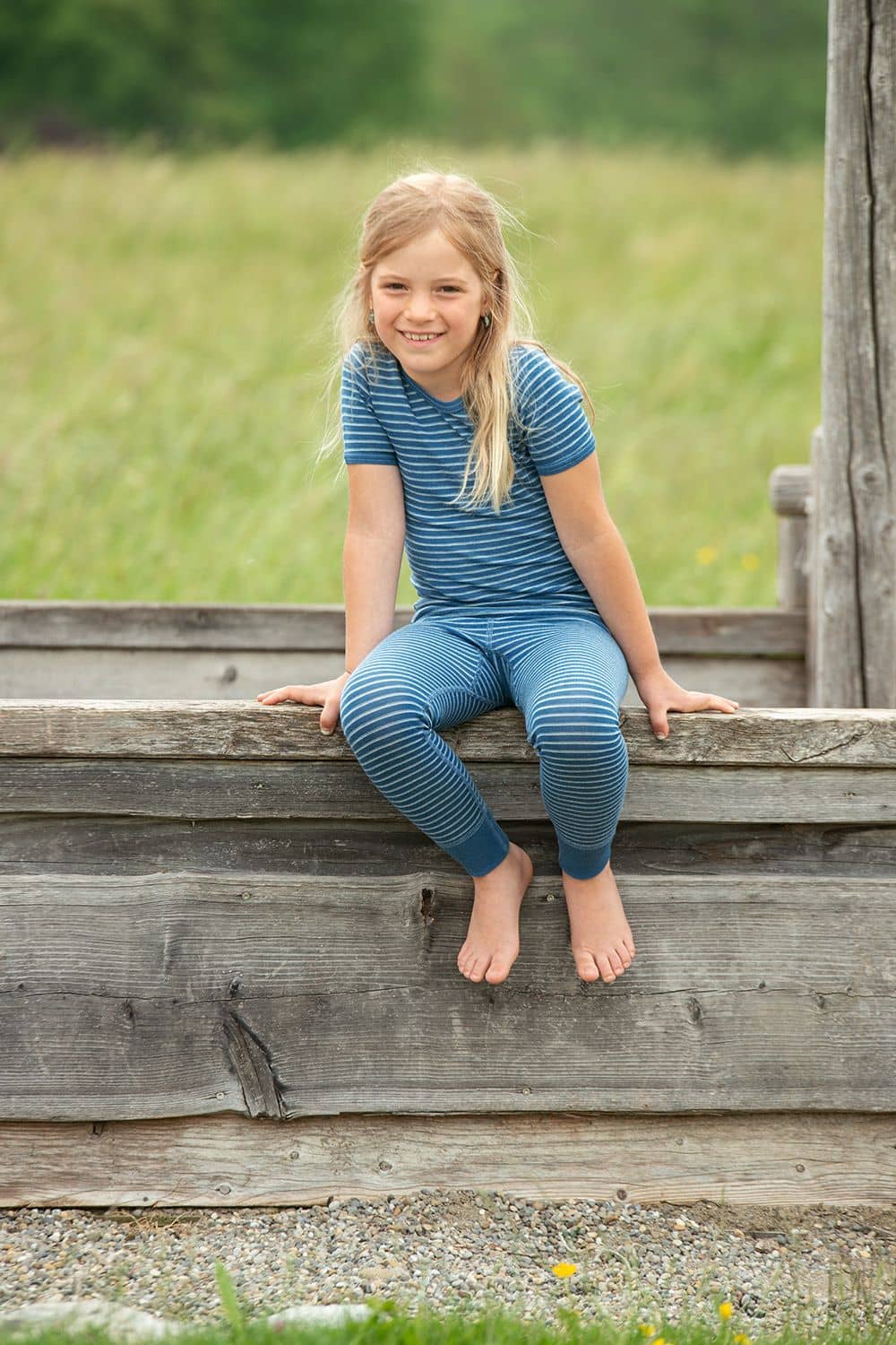 Geringeltes Wollhemd Kurzarm blau/naturweiß Mädchen sitzt in blau weiß geringeltes kurzarm Wollhemd aus bio Wolle (kbT) mit passender Streifenhose auf Holz