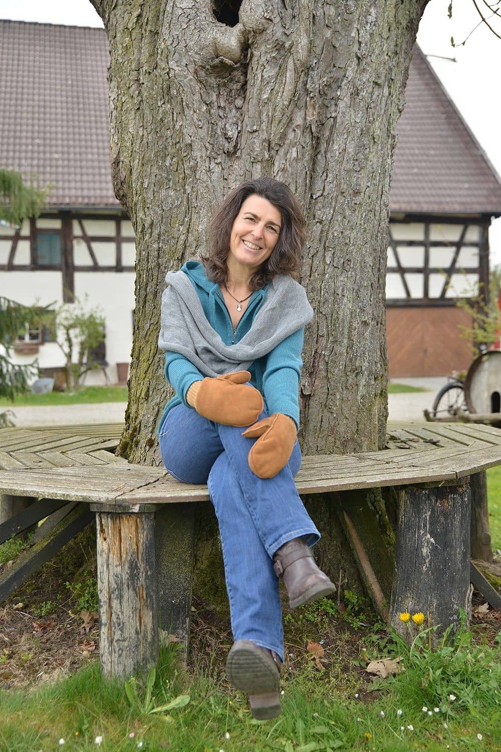 Frau sitzt auf Bank und trägt hellbraune Fell- Fausthandschuhe aus Pelzvelour mit Bund, ein blaues Shirt und grauen Schal