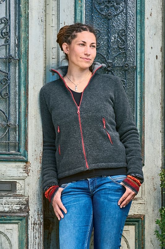 Frau lehnt an Holztür und trägt anthrazitfarbene Wollfleecejacke aus bio Wolle (kbT) mit Jeans und gemusterten Armstulpen