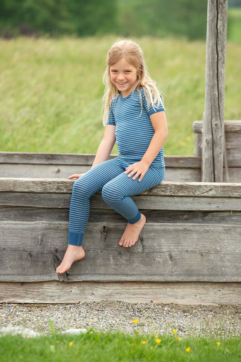 Mädchen sitzt mit blau weiß geringelte lange Wollunterhose aus bio Wolle (kbT) und passendem Ringelshirt auf Holz