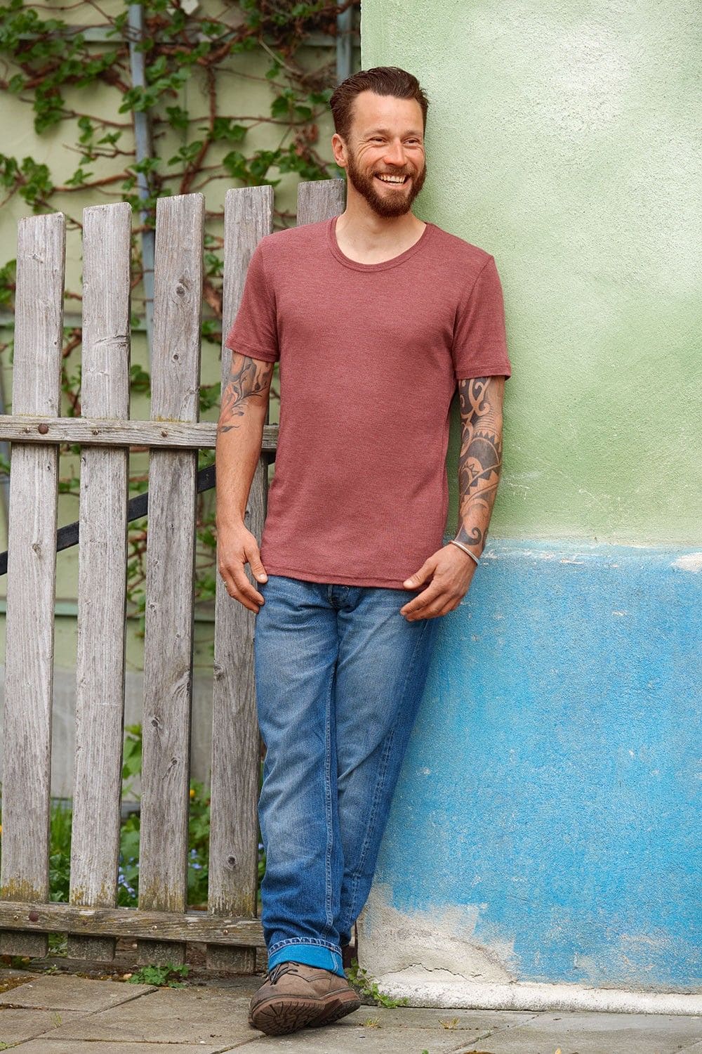 lachender Mann mit Jeans lehnt an Wand und trägt rostrotes Kurzarmshirt aus bio Wolle (kbT) und Seide