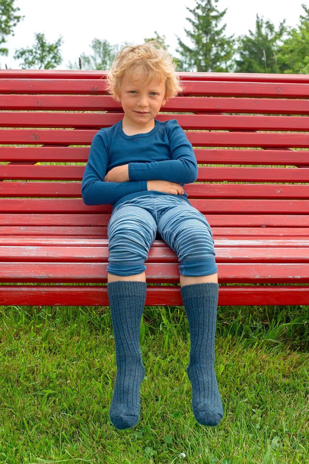 Junge sitzt auf roter Bank und trägt eine Jeans, ein blaues Shirt und blaumelierte Socken aus bio Wolle (kbT)