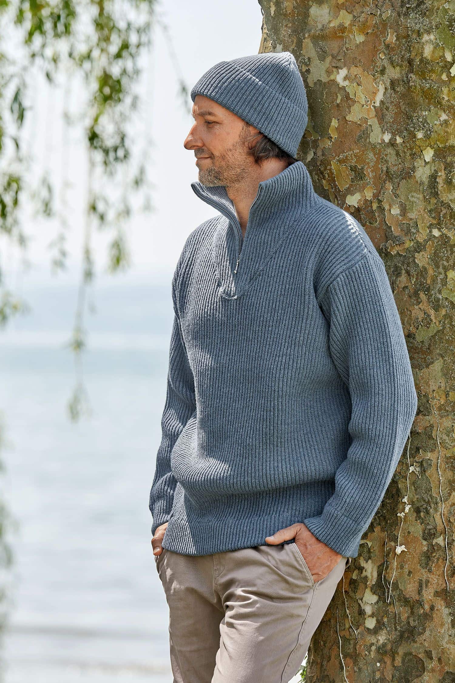 Mann lehnt sich an Baum und trägt eine rauchblaue Finkhof Mütze aus bio Wolle (kbT) und einen Troyer