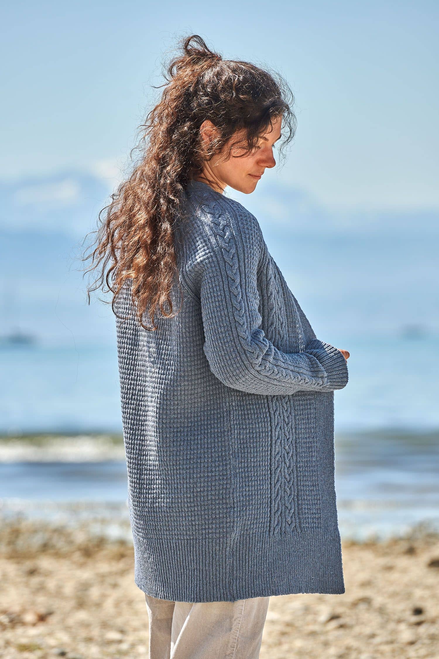 Rückansicht von Frau mit rauchblauem Cardigan mit Zopfmuster aus bio Wolle (kbT) vom Finkhof und beiger Jeans
