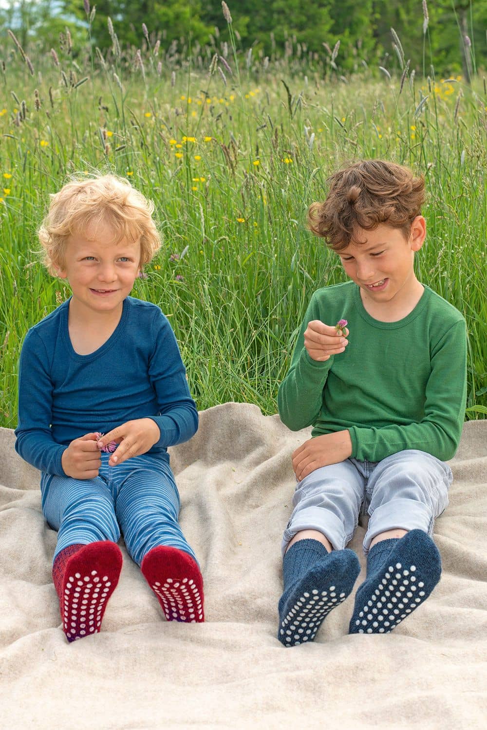 Wollplüsch-Stoppersocken Kinder sitzen mit roten und blauen Stoppersocken aus bio Wolle (kbT) auf Decke und spielen mit Blumen