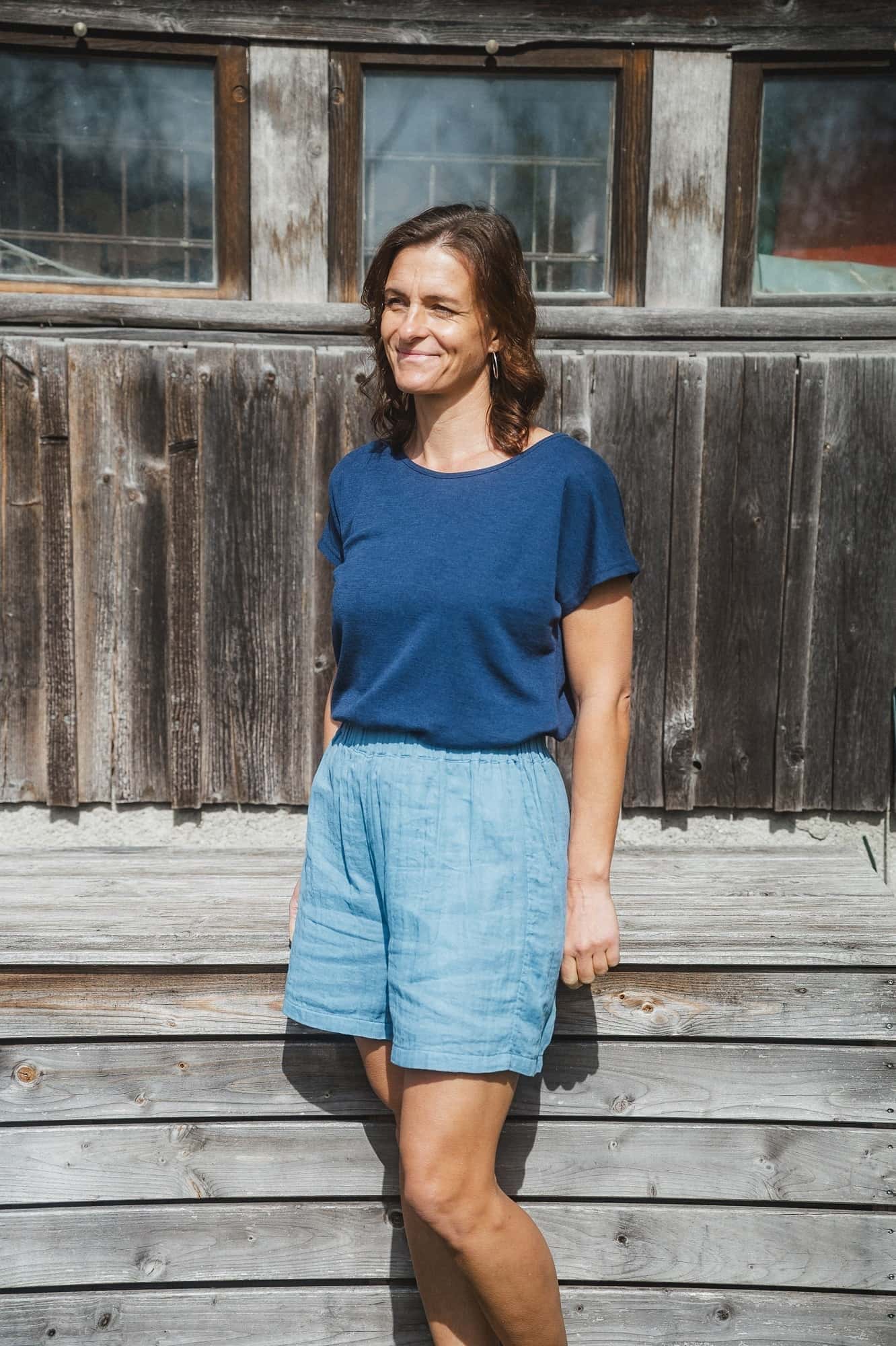 Frau steht vor Holzwand und trägt ein blaues Shirt und eine blaue Shorts aus Bio-Baumwoll-Musselin (kbA) vom Finkhof
