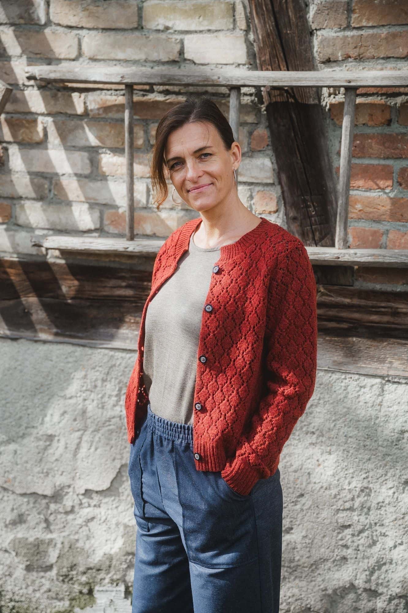 Frau trägt eine weite Hose und ein braunes Shirt mit einer rot orangen Strickjacke mit Ajourmuster aus Bio-Wolle vom Finkhof