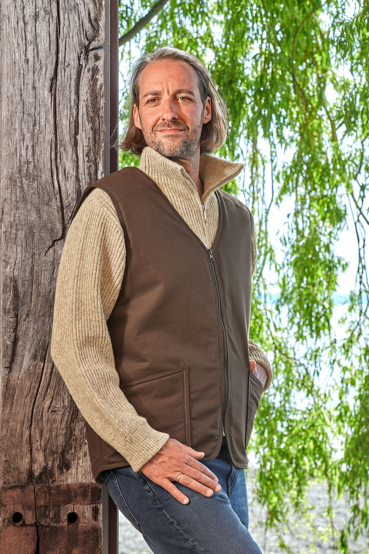 Mann lehnt sich an Baum und trägt einen Troyer und die braune Fellweste aus bio Baumwolle (kbA) und Fell vom Finkhof