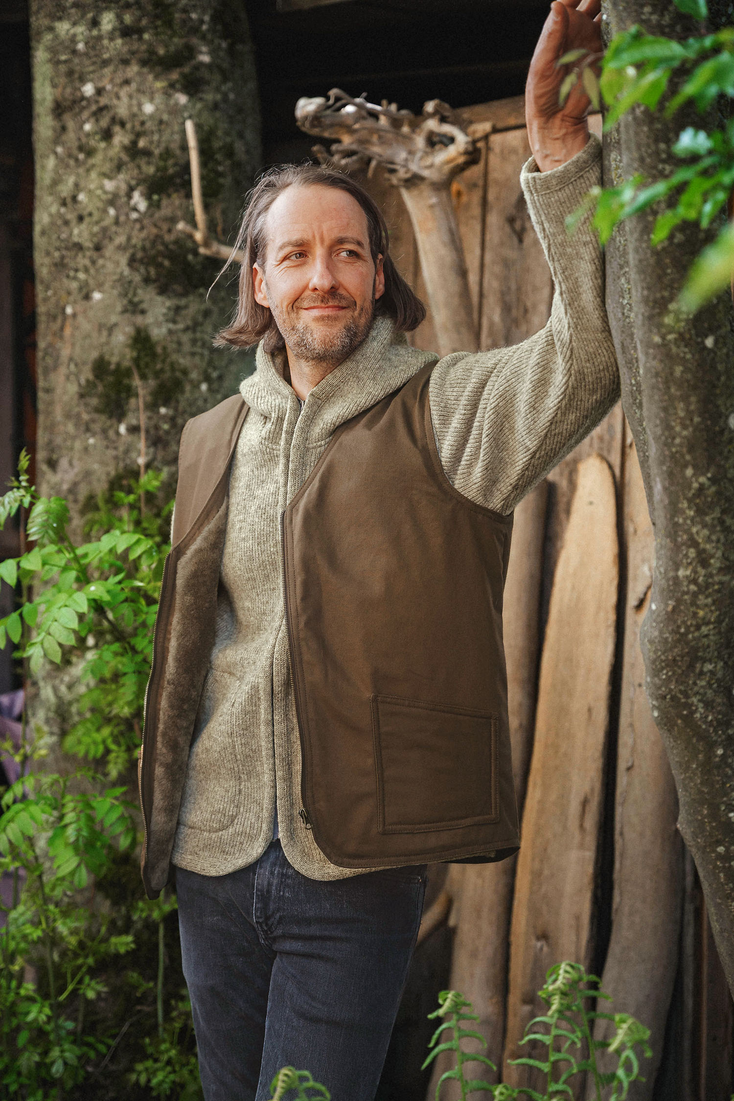 Mann trägt braune Fellweste aus bio Baumwolle (kbA) und Fell vom Finkhof im Wald mit grauer Jacke