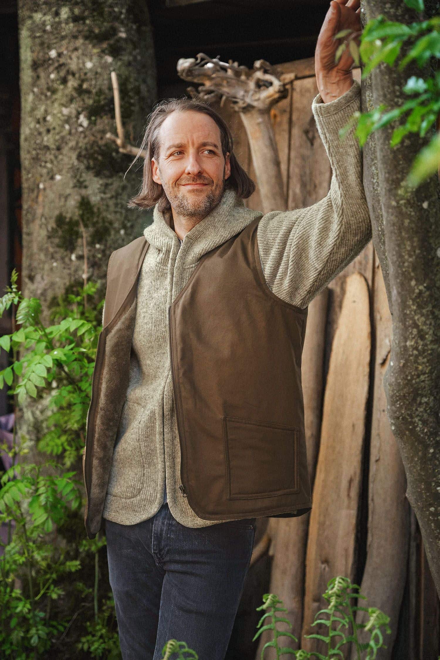 Herren-Fellweste braun Gr. M Mann trägt braune Fellweste aus bio Baumwolle (kbA) und Fell vom Finkhof im Wald mit grauer Jacke