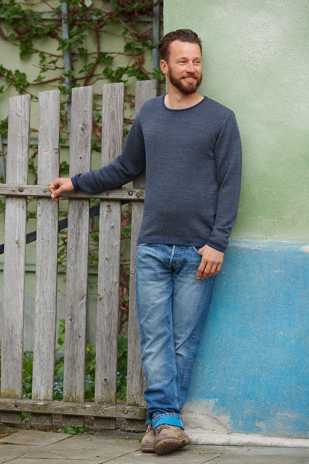 Mann lehnt an grüner Wand und trägt blauen Melangepullover aus bio Wolle (kbT) vom Finkhof