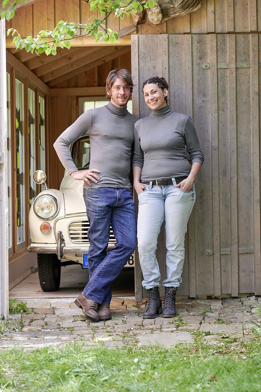 Mann und Frau stehen vor Holztor und tragen graue Pullover mit Rollkragen aus bio Baumwolle (kbA) und Seide