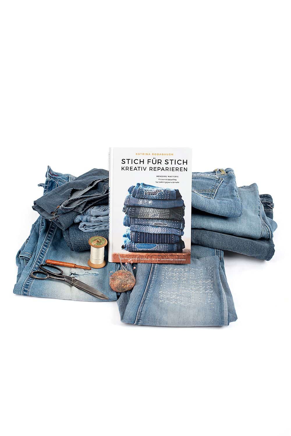 Stich für Stich kreativ reparieren Buch ,,Stich für Stich kreativ reparieren" angelehnt an Stapel Jeanshosen und Jeansstücke sowie Zubehör im Vordergrund