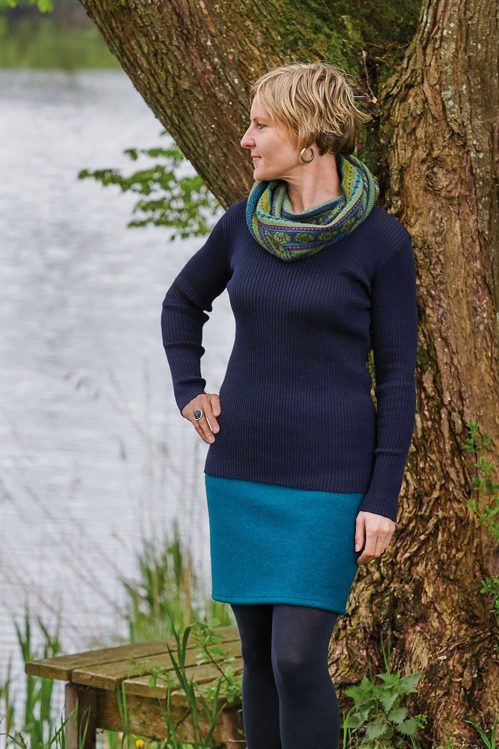 Frau vor einem Baum trägt blaues Rippshirt, türkiser Rock und grün gemusterter Loopschal aus bio Wolle (kbT)