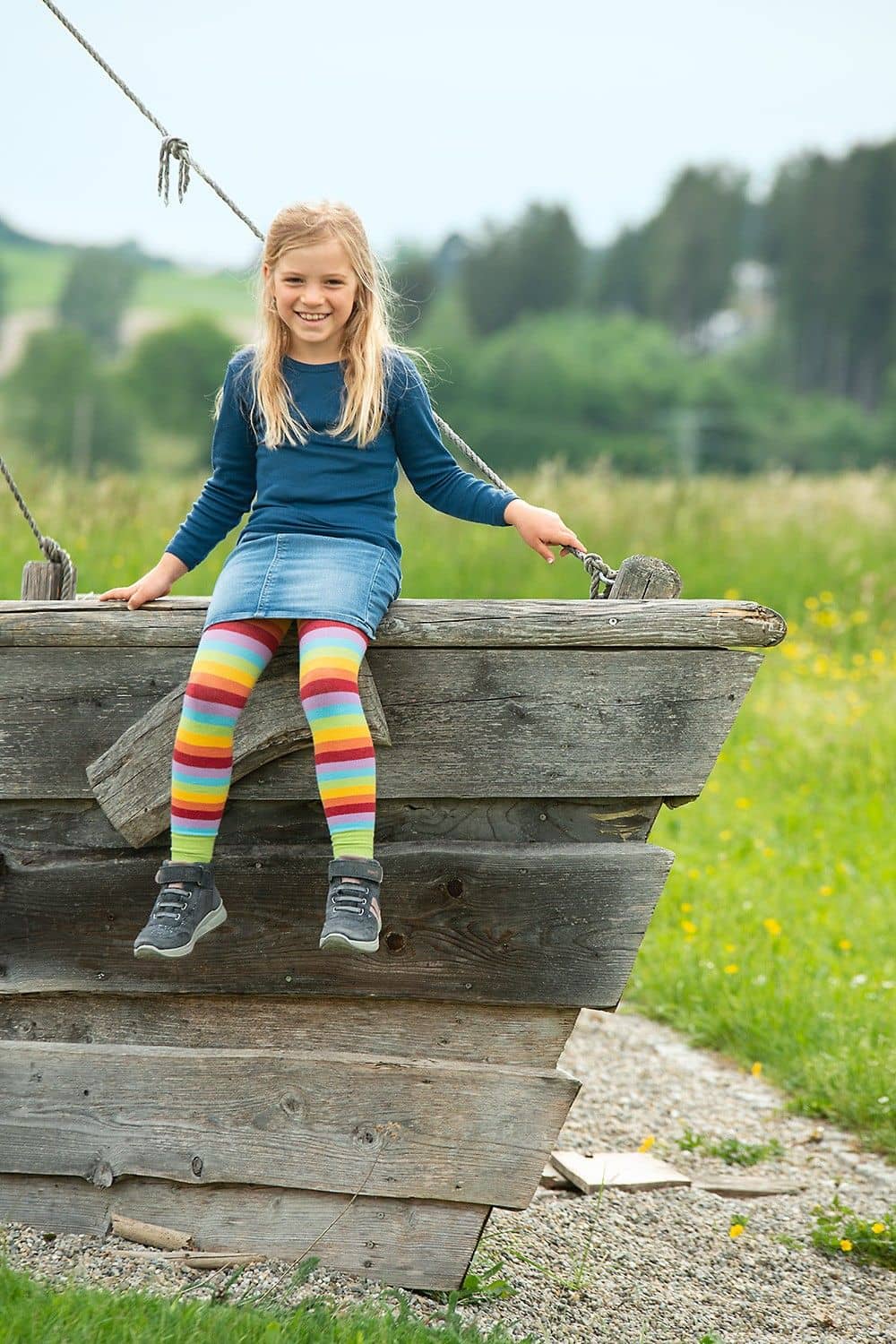 Kind sitzt auf einem Holzschiff und trägt eine bunte gestreifte Leggings aus bio Baumwolle (kbA), Elasthan mit Jeansrock