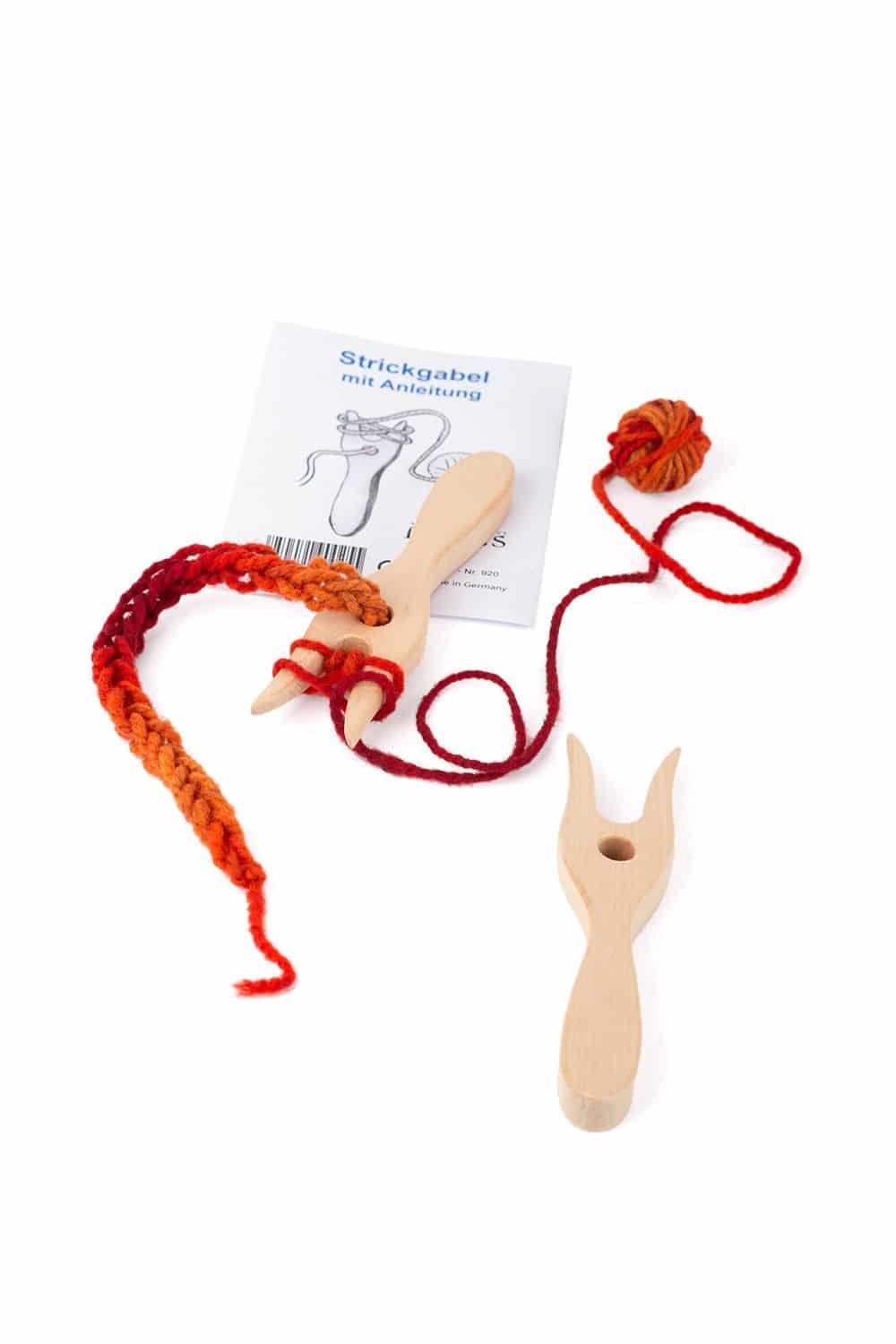 Holzstrickgabel liegt auf Anleitung mit Strickstück aus rot oranger Wolle, zusätzlich liegt noch eine weitere Strickgabel im Vordergrund