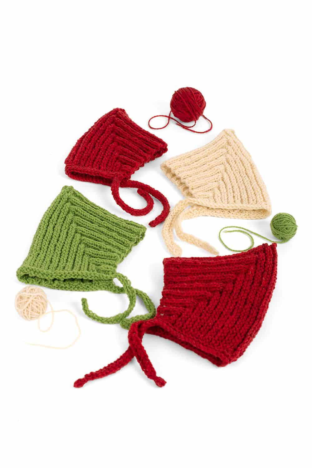 weiße, rote und grüne Zwergenmützen liegen mit kleinen Wollknäulen auf weißem Untergrund für Strickanleitung