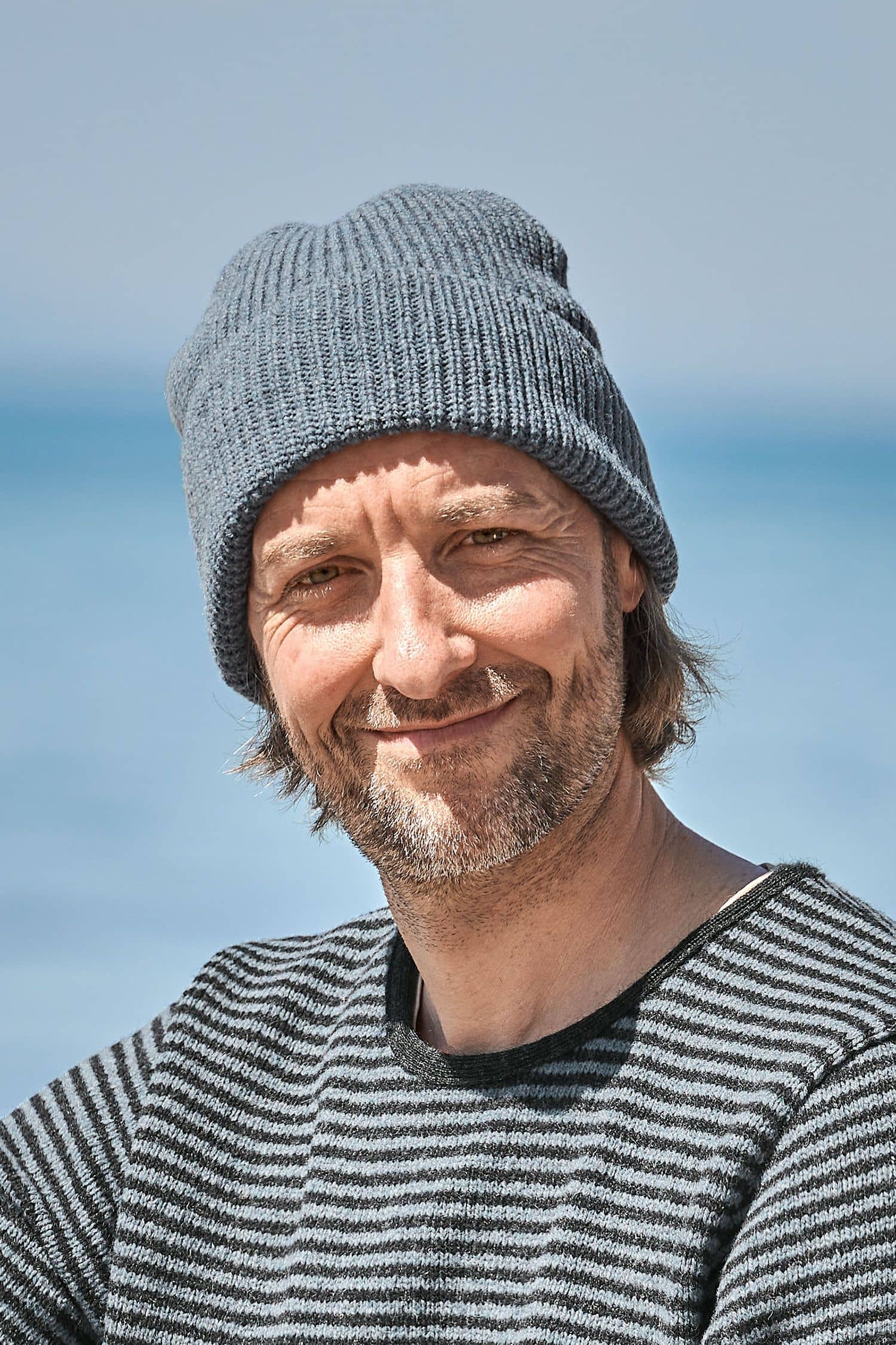 Mann trägt Streifenpulli und eine rauchblaue Finkhof Mütze aus bio Wolle (kbT)