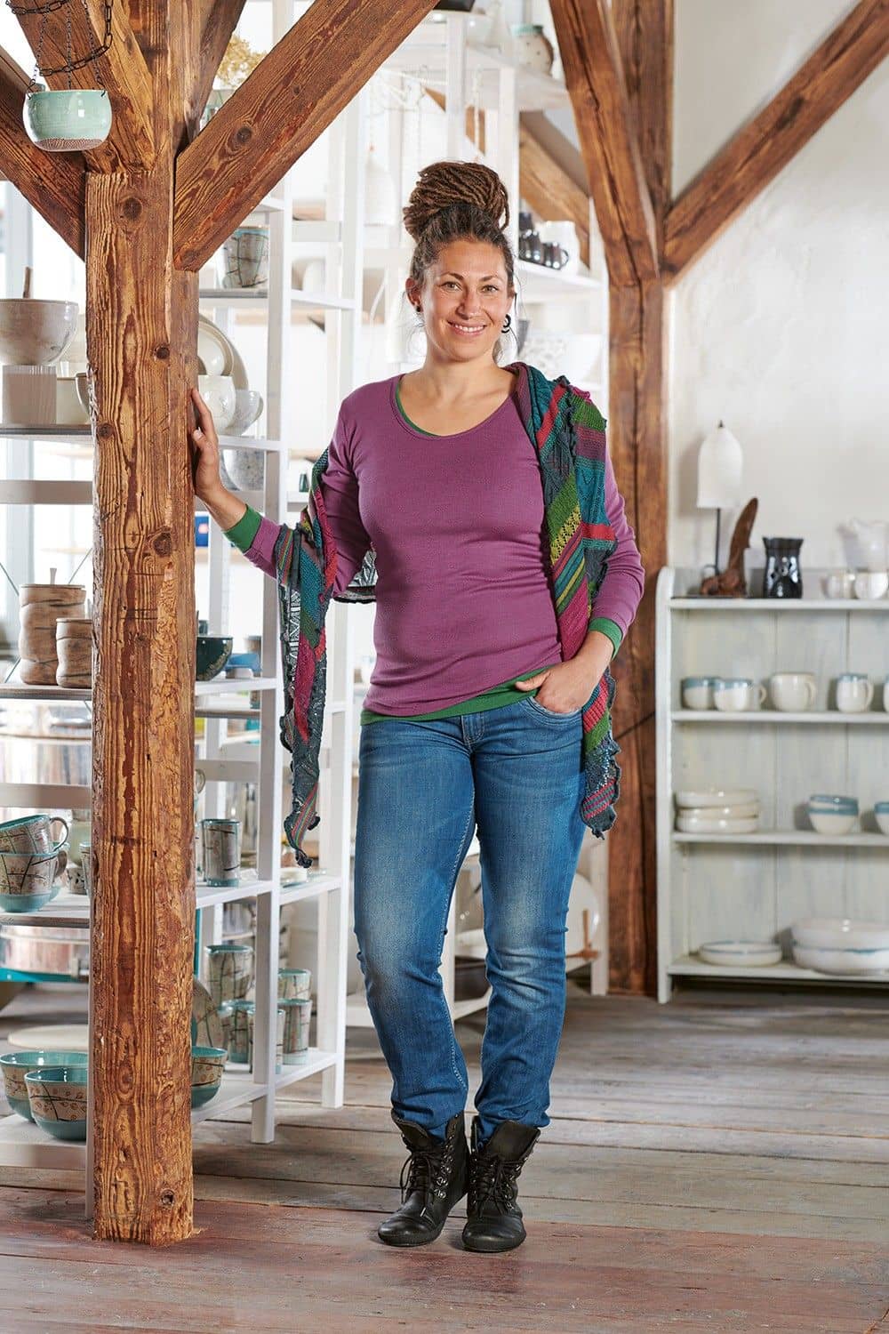 Frau steht neben Regalen mit Geschirr und trägt Jeans, buntes Tuch und lila Langarmshirt aus bio Wolle (kbT) und Seide