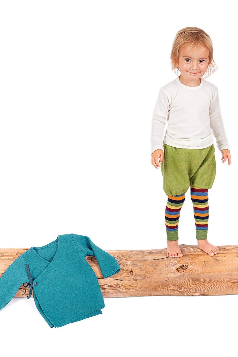 Kind steht aus Balken und trägt Shirt, gestreifte Hose und grüne kurze Hose, daneben liegt Pulli aus petrolblauem Strickwalk