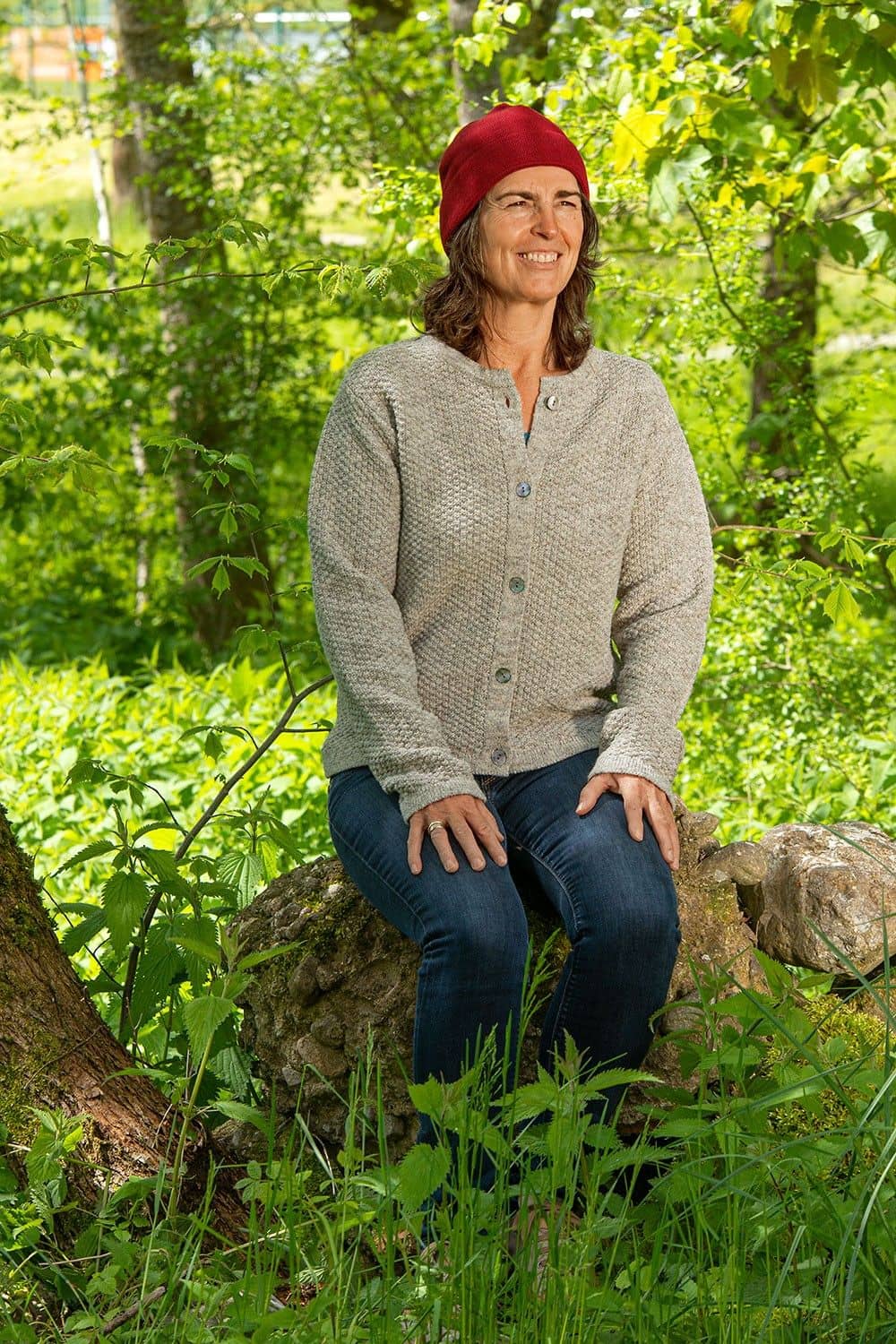 Frau sitzt unter Bäumen auf einem Stein und trägt rote Mütze, Jeans und graue Finkhof Strukturjacke aus bio Wolle (kbT)
