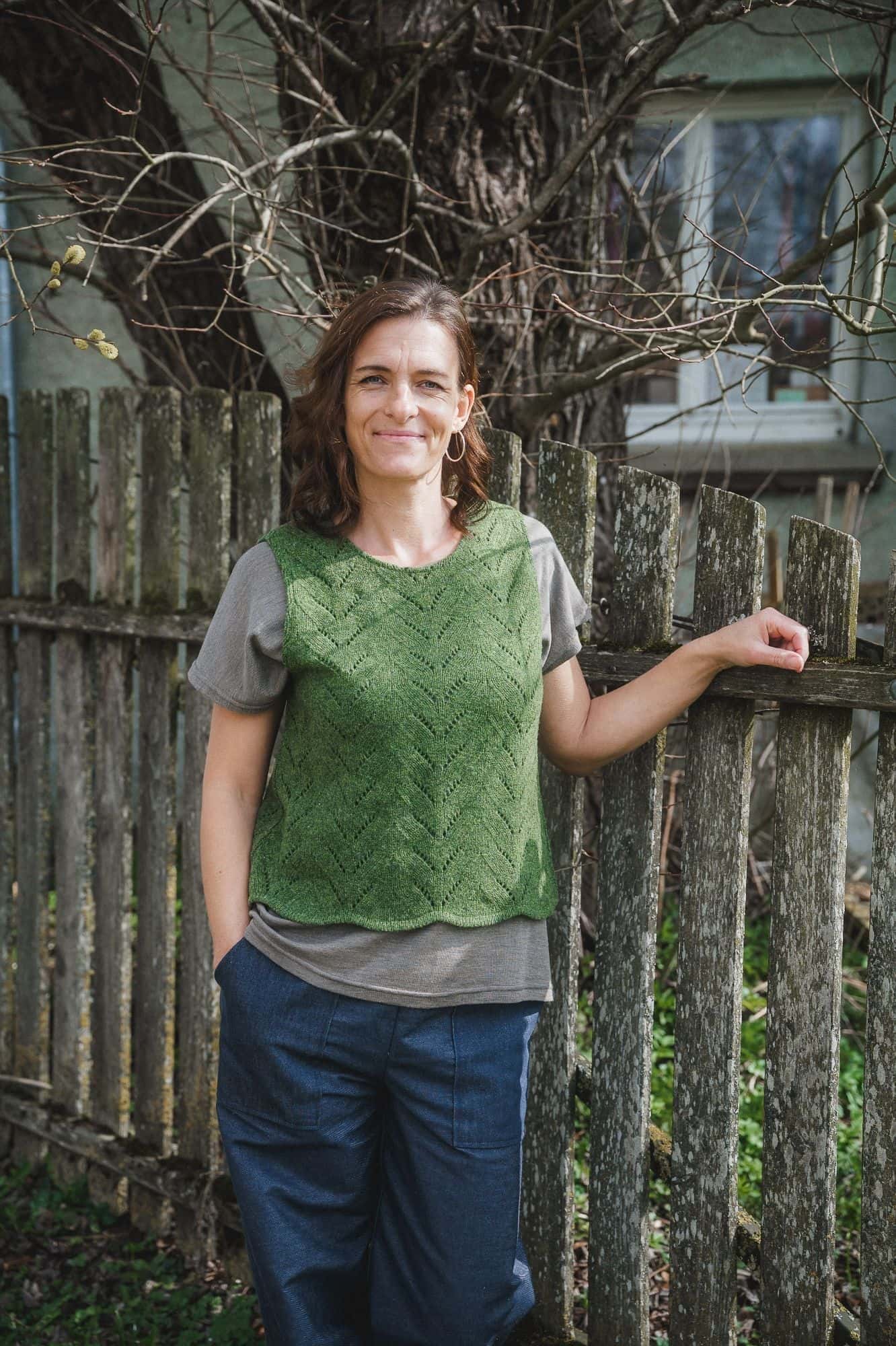 Frau steht am Zaun und trägt ein weites Shirt mit einem grünen Strickpullunder mit Ajourmuster aus Bio-Wolle vom Finkhof