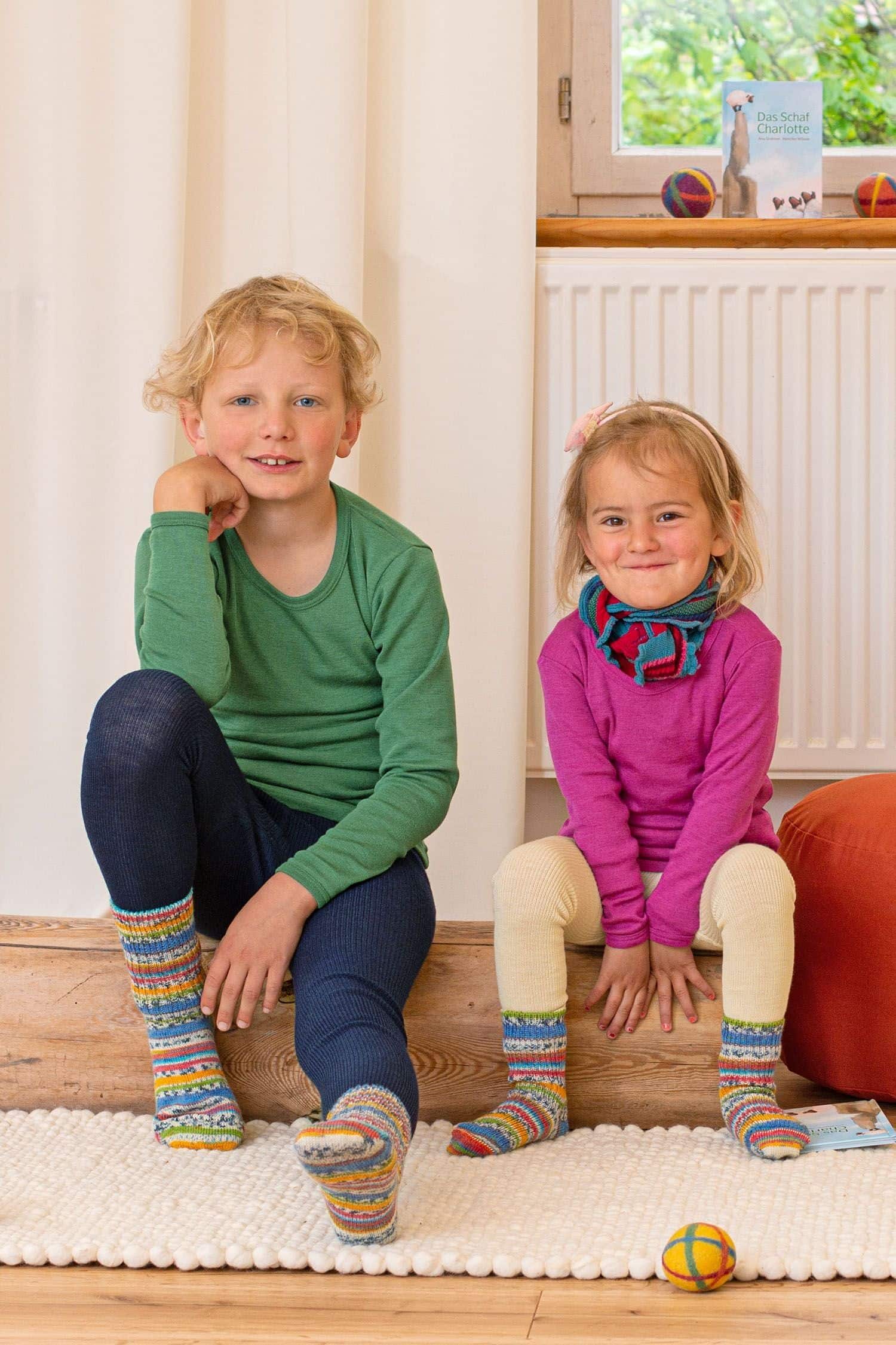 Wollsocken Kunterbunt Kinder mit Wollkleidung sitzen auf einem Holzbalken und tragen bunte Wollsocken