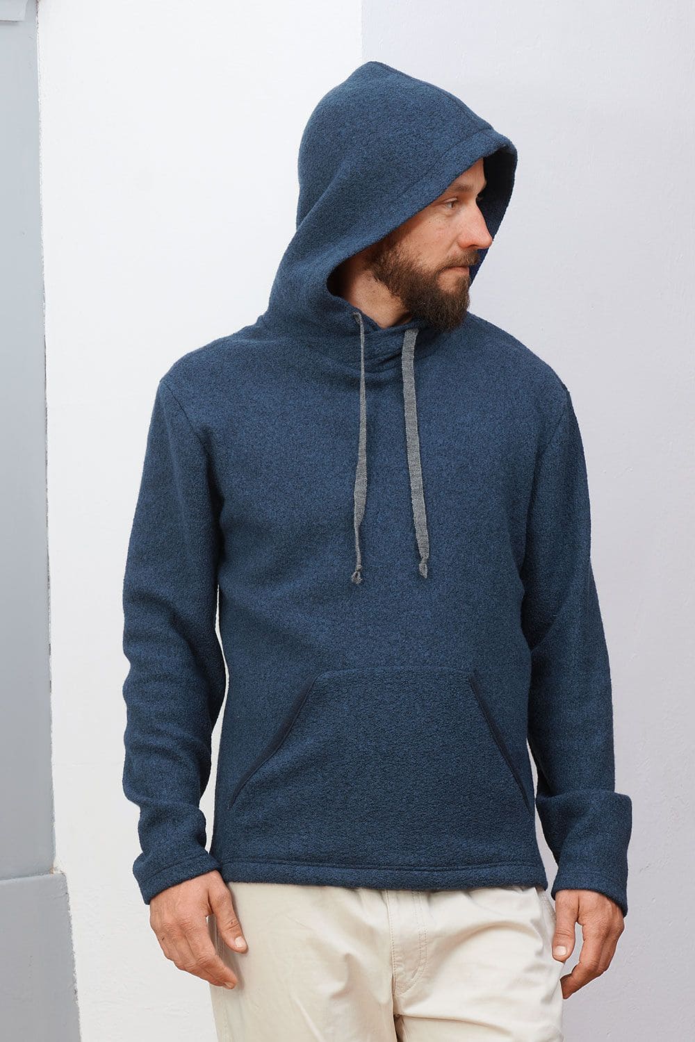 Mann steht vor Wand und trägt marineblauen Kapuzenpullover aus bio Wolle (kbT) und hat Kapuze auf dem Kopf