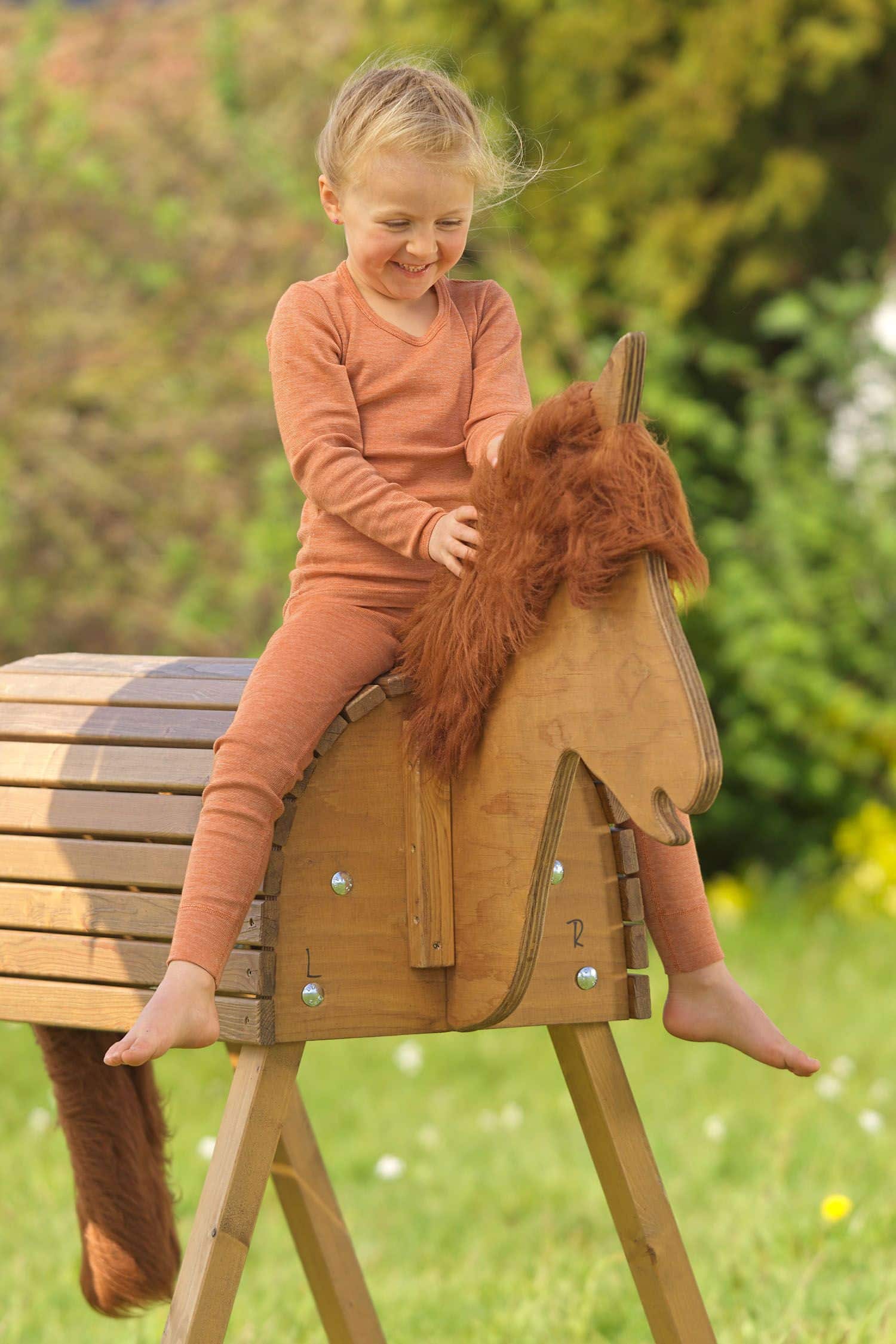 Kind sitzt auf Holzpferd und trägt eine orange lange Unterhose aus bio Wolle (kbT), bio Baumwolle (kbA) und Seide mit Shirt
