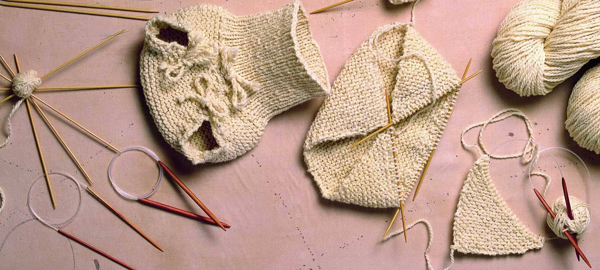 Strickanleitung für Windelhosen fertige und angefangene Windelhose mit Wollen und Stricknadeln auf rosa Untergrund für Strickanleitung